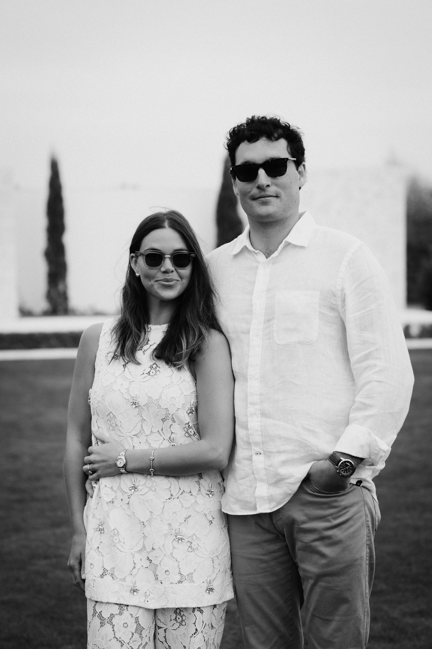 A couple in sunglasses and coordinating white outfits posing outdoors.