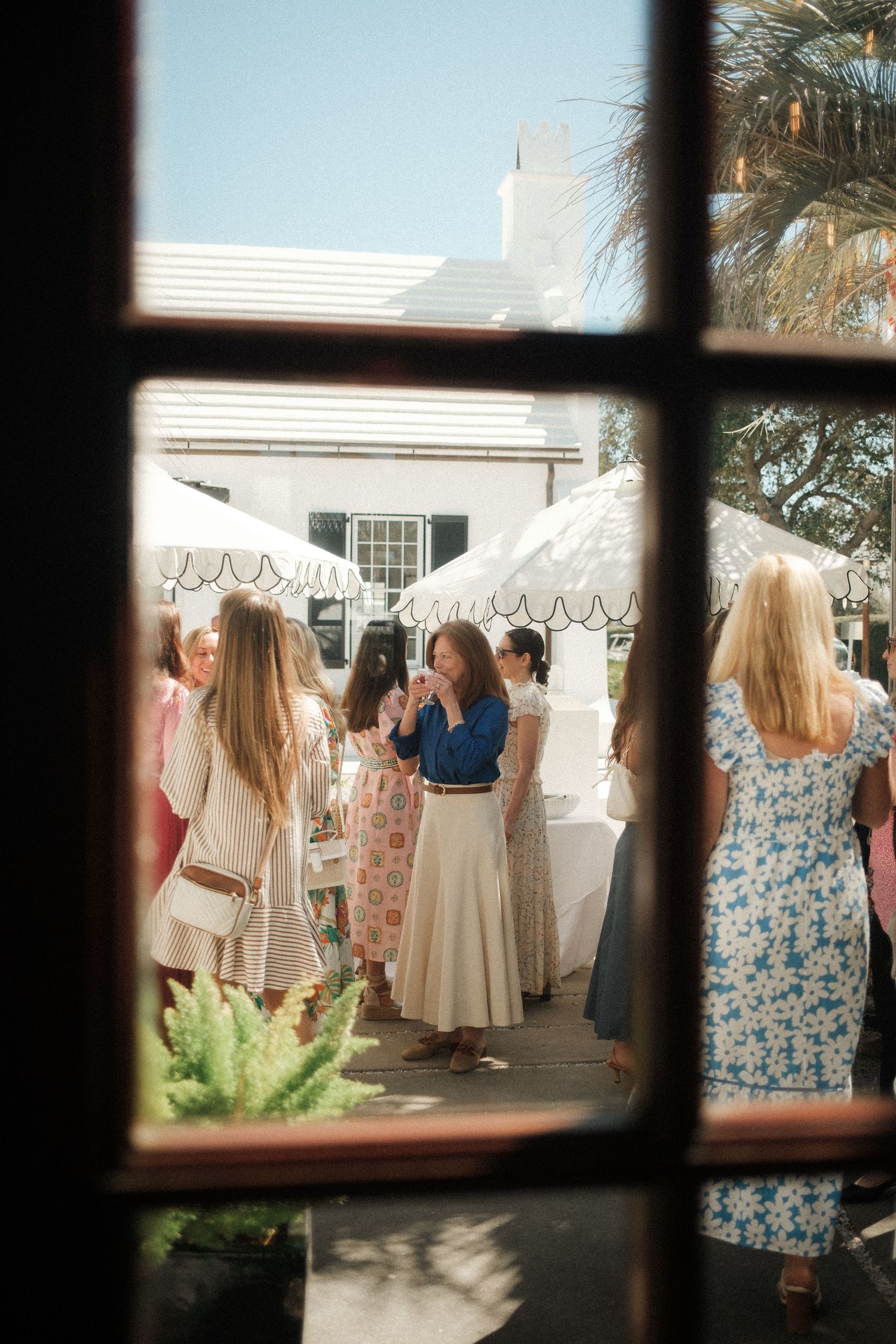 Women in summer dresses gather outdoors at a garden market event, viewed through a wooden window frame on a sunny day.