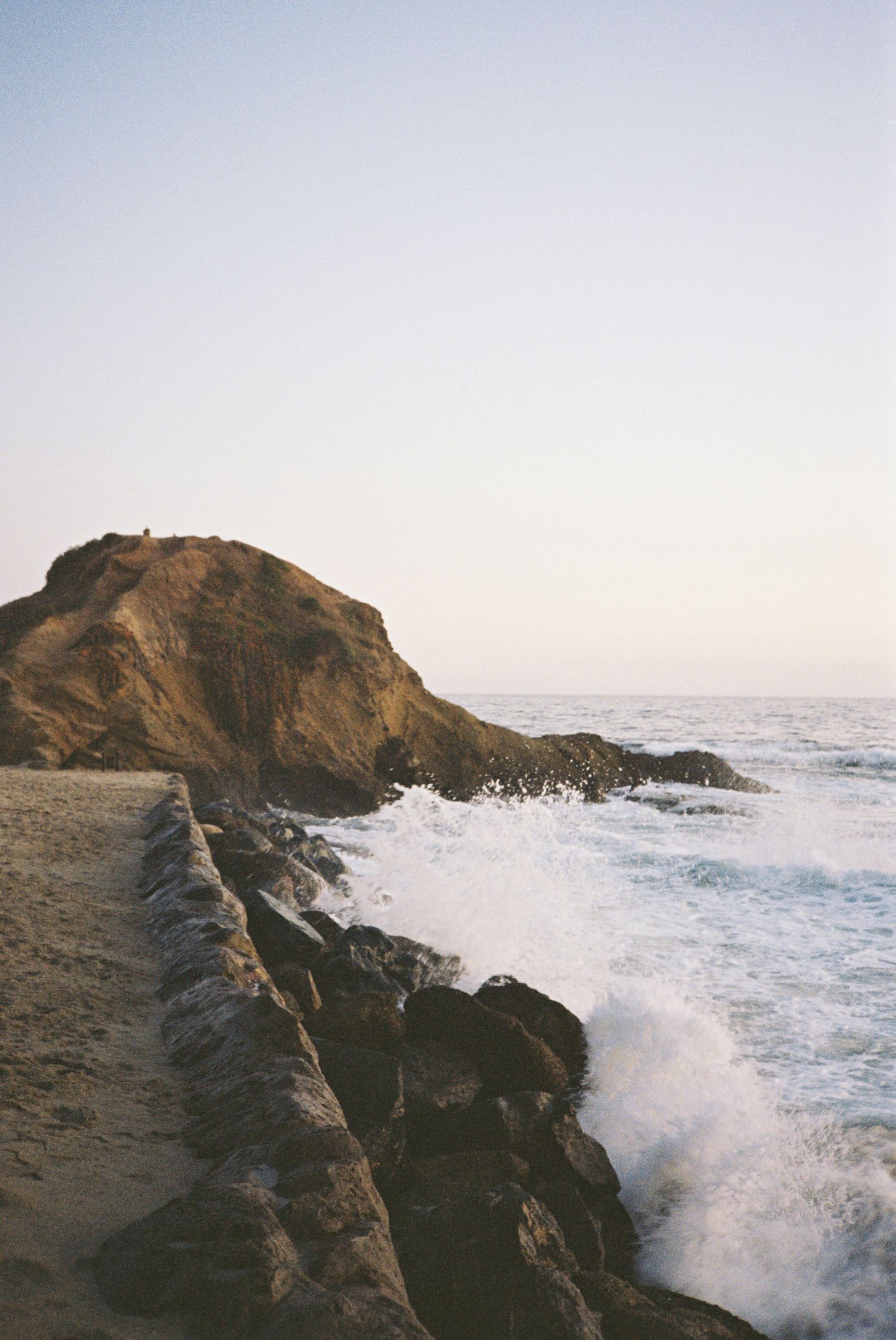 Ocean waves crash dramatically against rocky coastal cliffs during sunset creating a misty spray against the rugged shore.