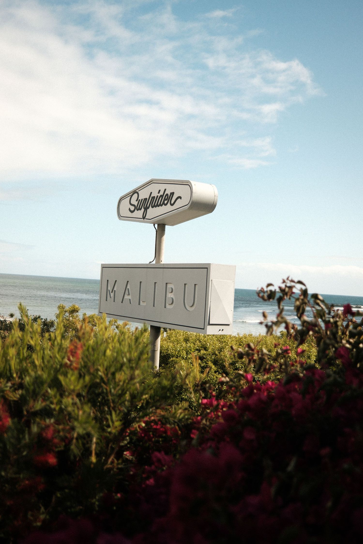 A vintage-style Malibu sign stands against a blue sky with white clouds, framed by green foliage and red flowers.