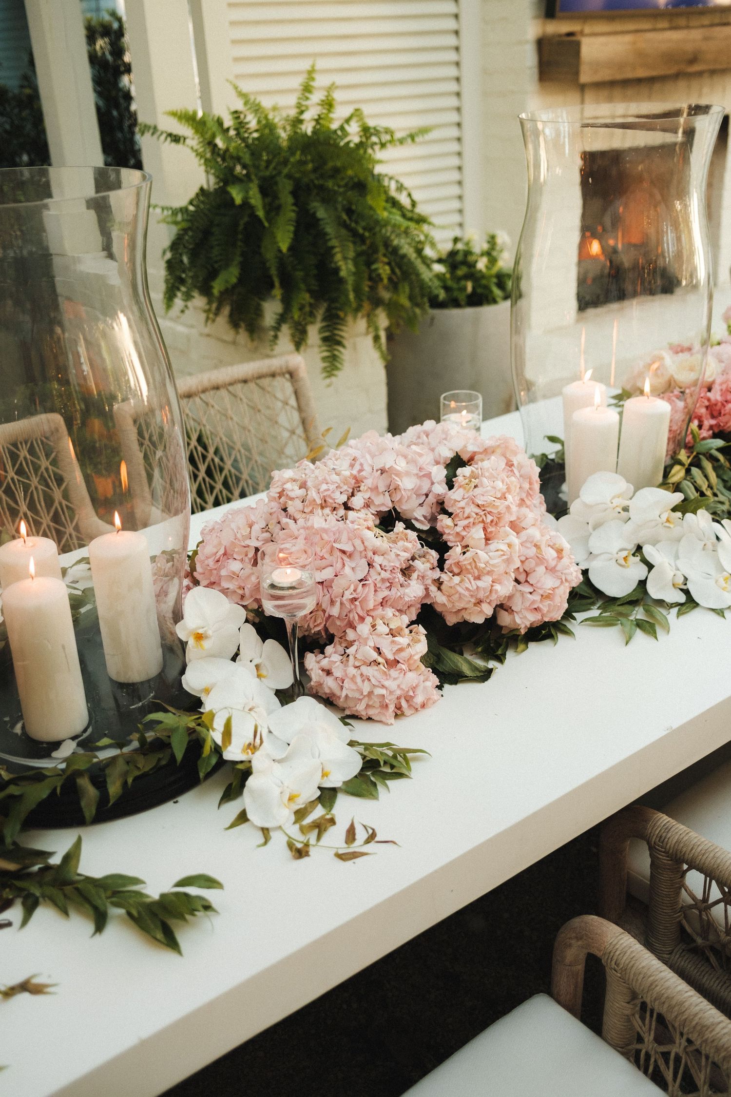 Elegant wedding table with pink hydrangeas, white orchids, greenery, and lit pillar candles on a white surface.