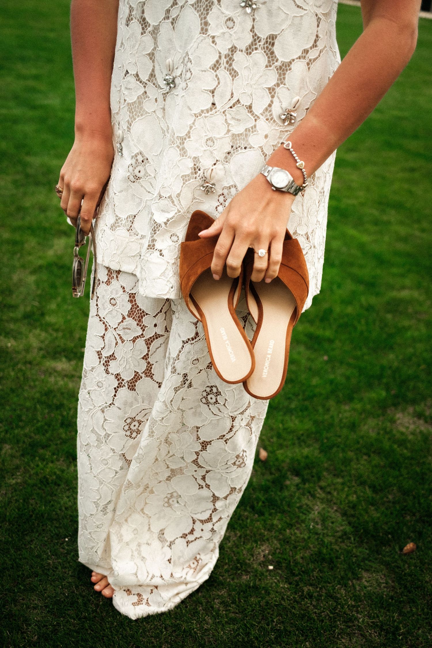 Close-up of hands holding brown sandals against a white lace dress on grass.