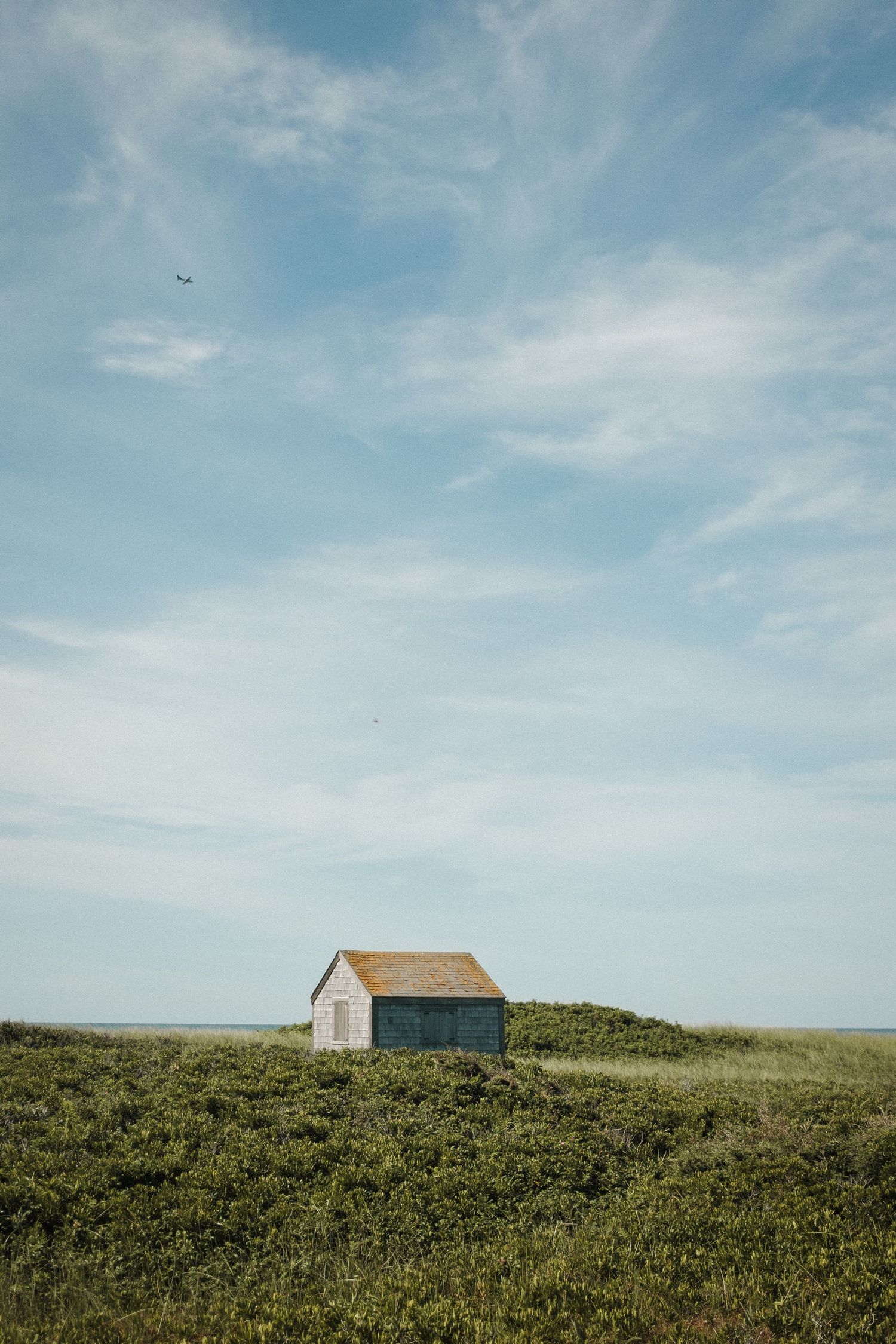 Two small houses stand alone on a grassy hill against a bright blue sky with wispy clouds.