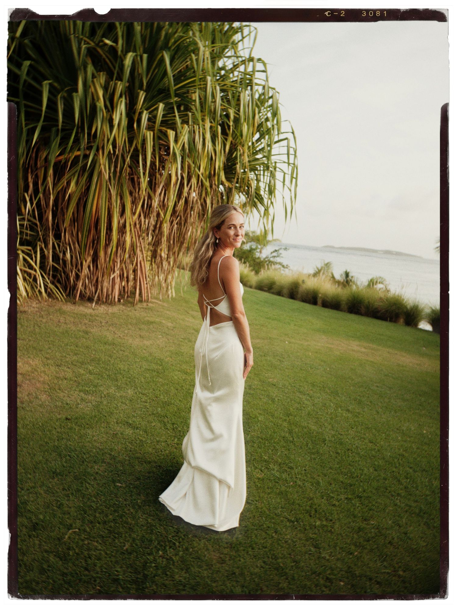 Woman in elegant white backless gown standing on lush green lawn with tropical palms and ocean view.