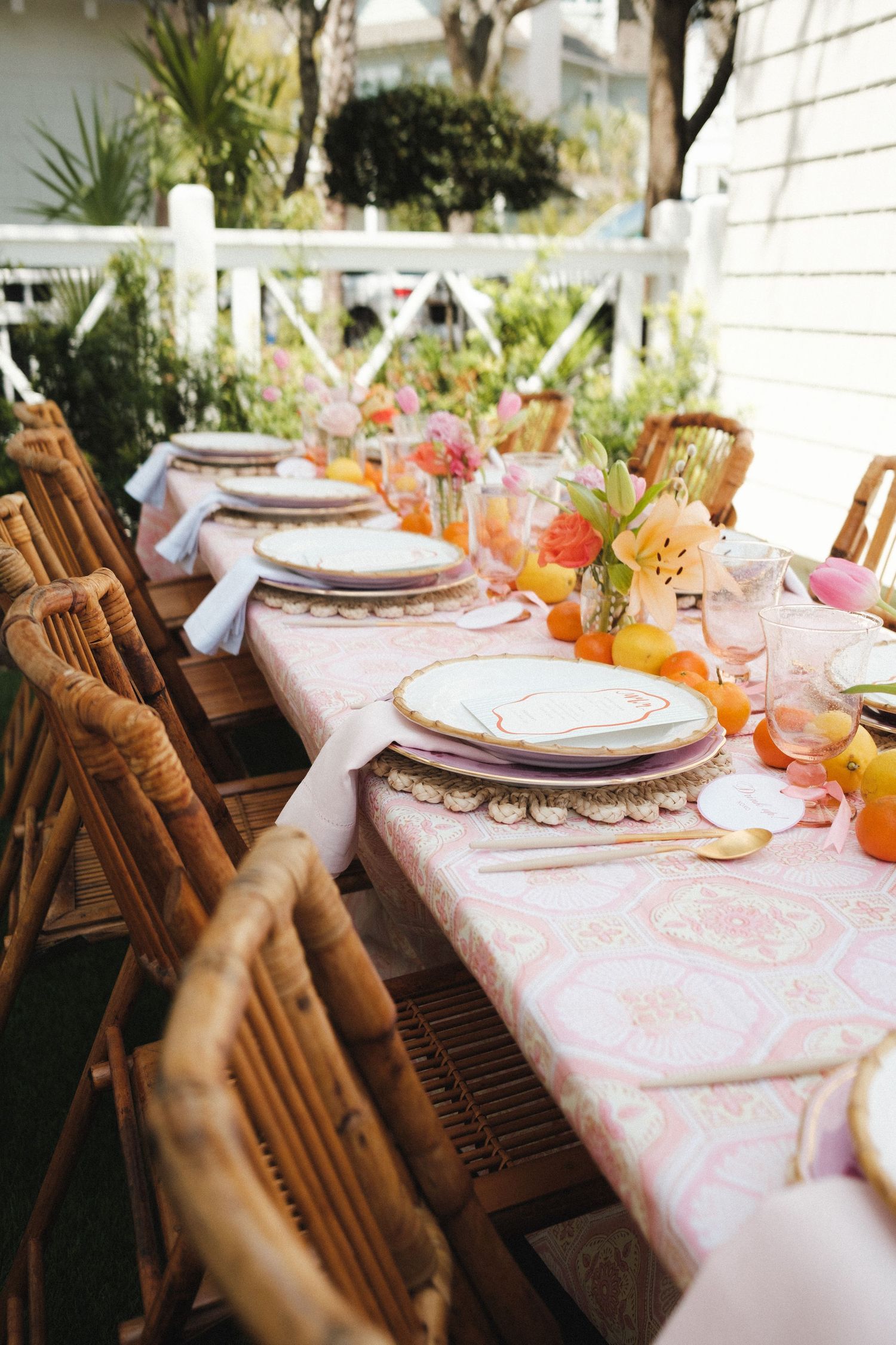 Elegant outdoor dining table with pink floral tablecloth, wicker chairs, gold-rimmed plates, and colorful spring flower centerpieces.