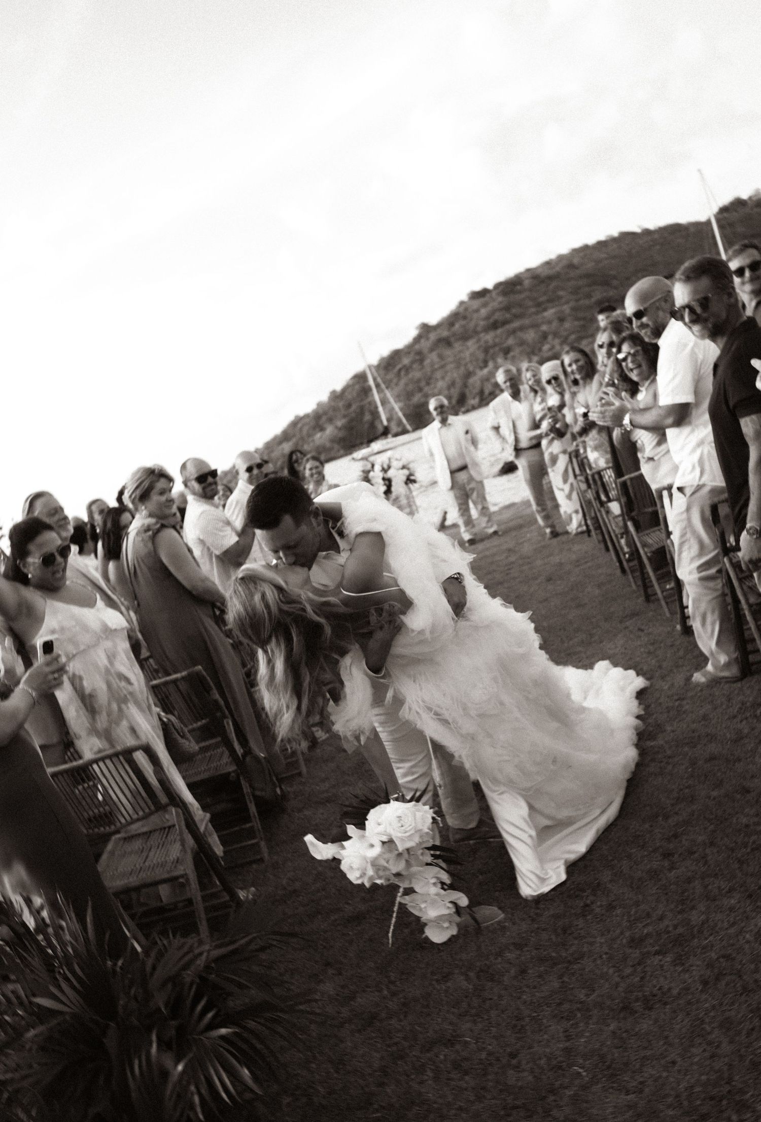 Black and white outdoor wedding ceremony with bride and groom kissing along the aisle surrounded by seated guests.