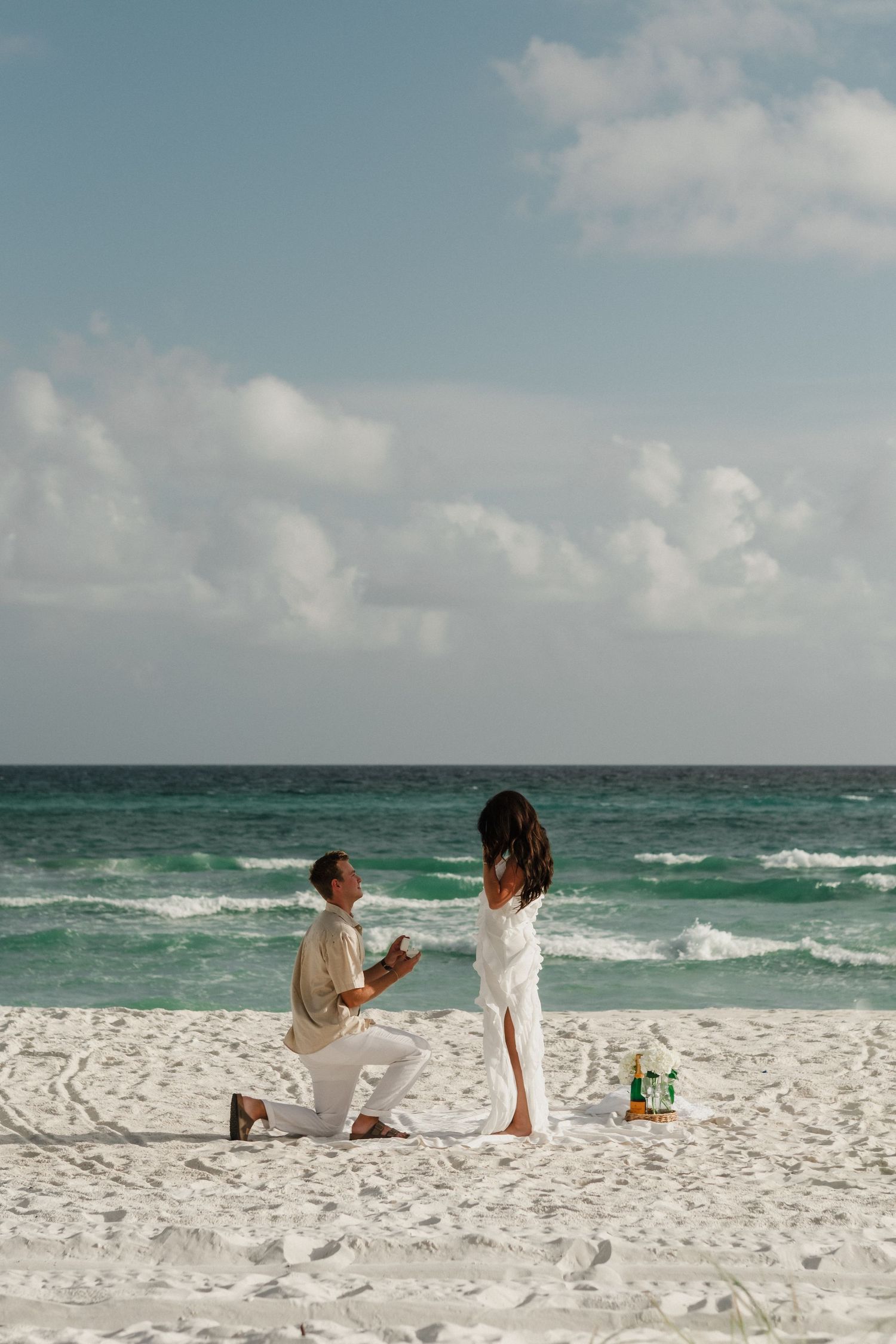 A romantic beach proposal scene with two figures kneeling on white sand as turquoise waves crash in the background.