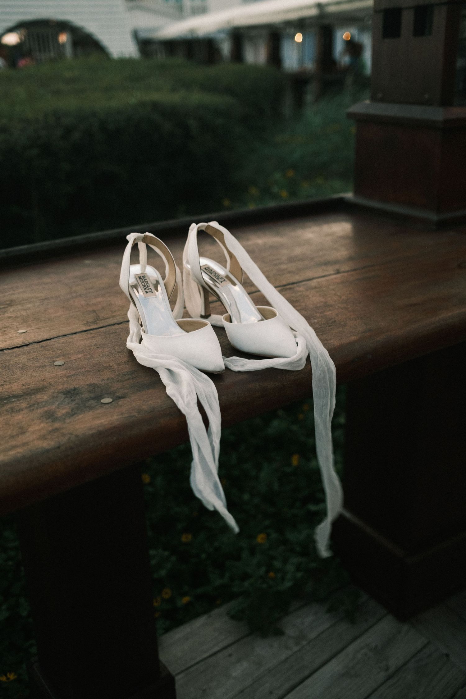 White bridal heels with long ribbon ties resting on a wooden deck railing with lush green hedges in the background.