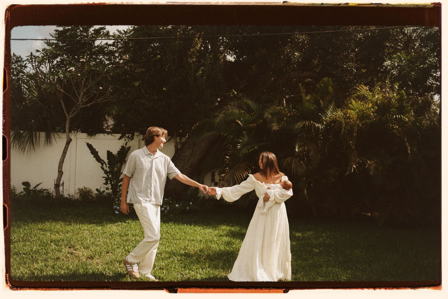A couple in white outfits hold hands and dance together on a green lawn surrounded by lush tropical trees.