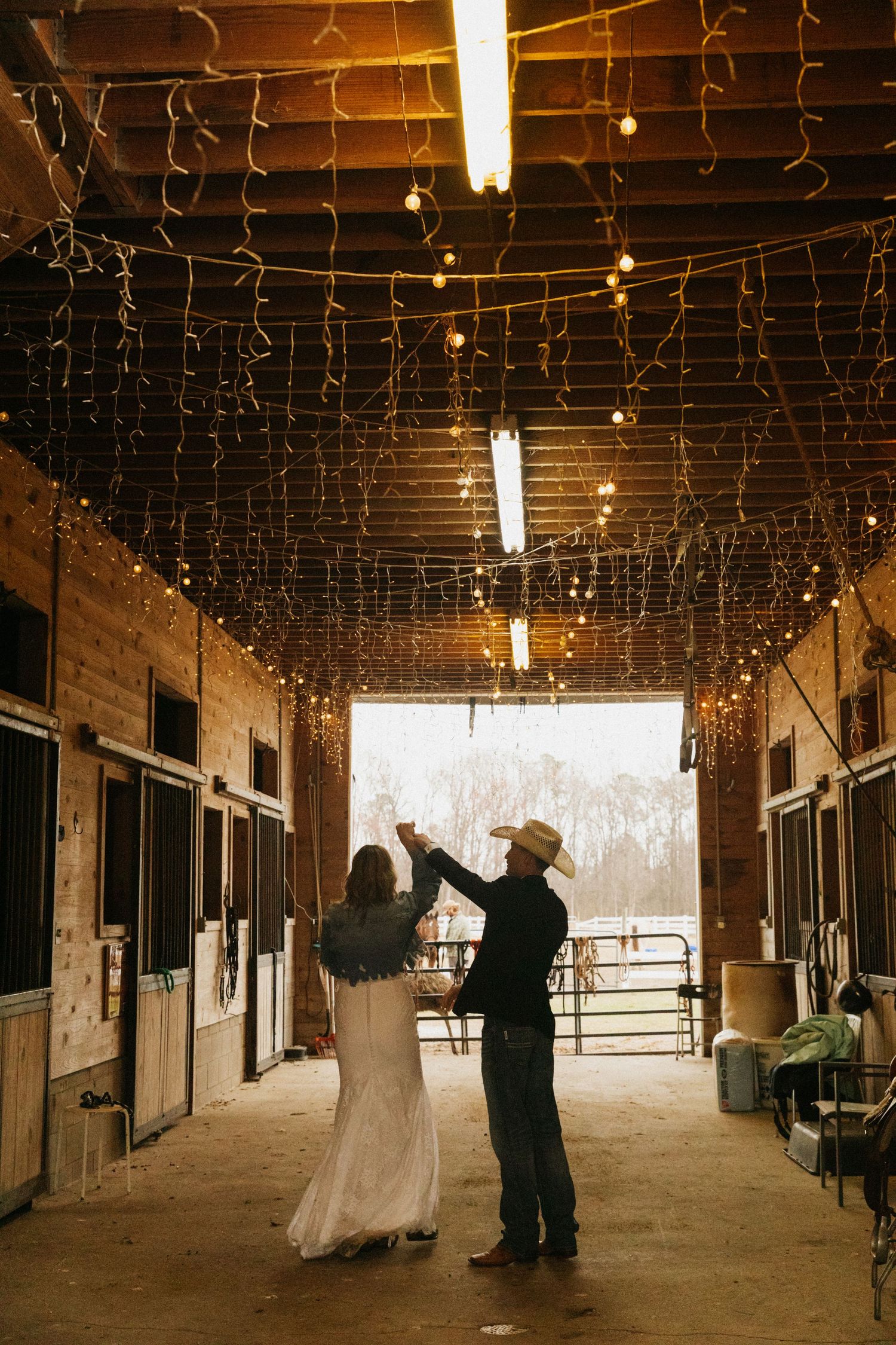 Couple dancing in a rustic barn adorned with string lights, wearing wedding attire and cowboy hat.