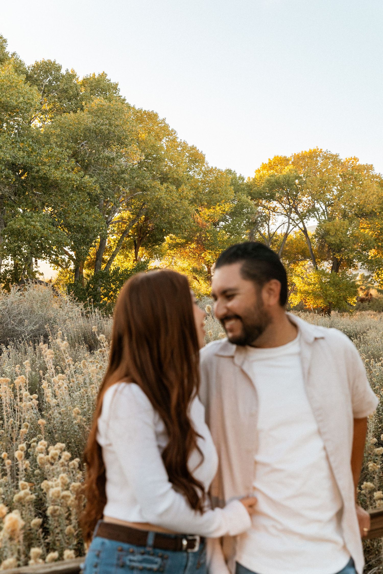 Couple smiling at each other in a golden hour field with wildflowers and autumn trees in the background.