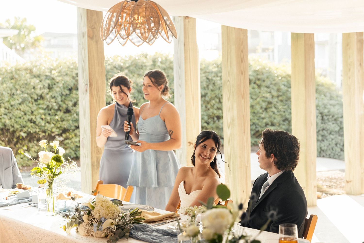 Bridesmaids in blue dresses give a toast speech at an elegant wedding reception with floral centerpieces.