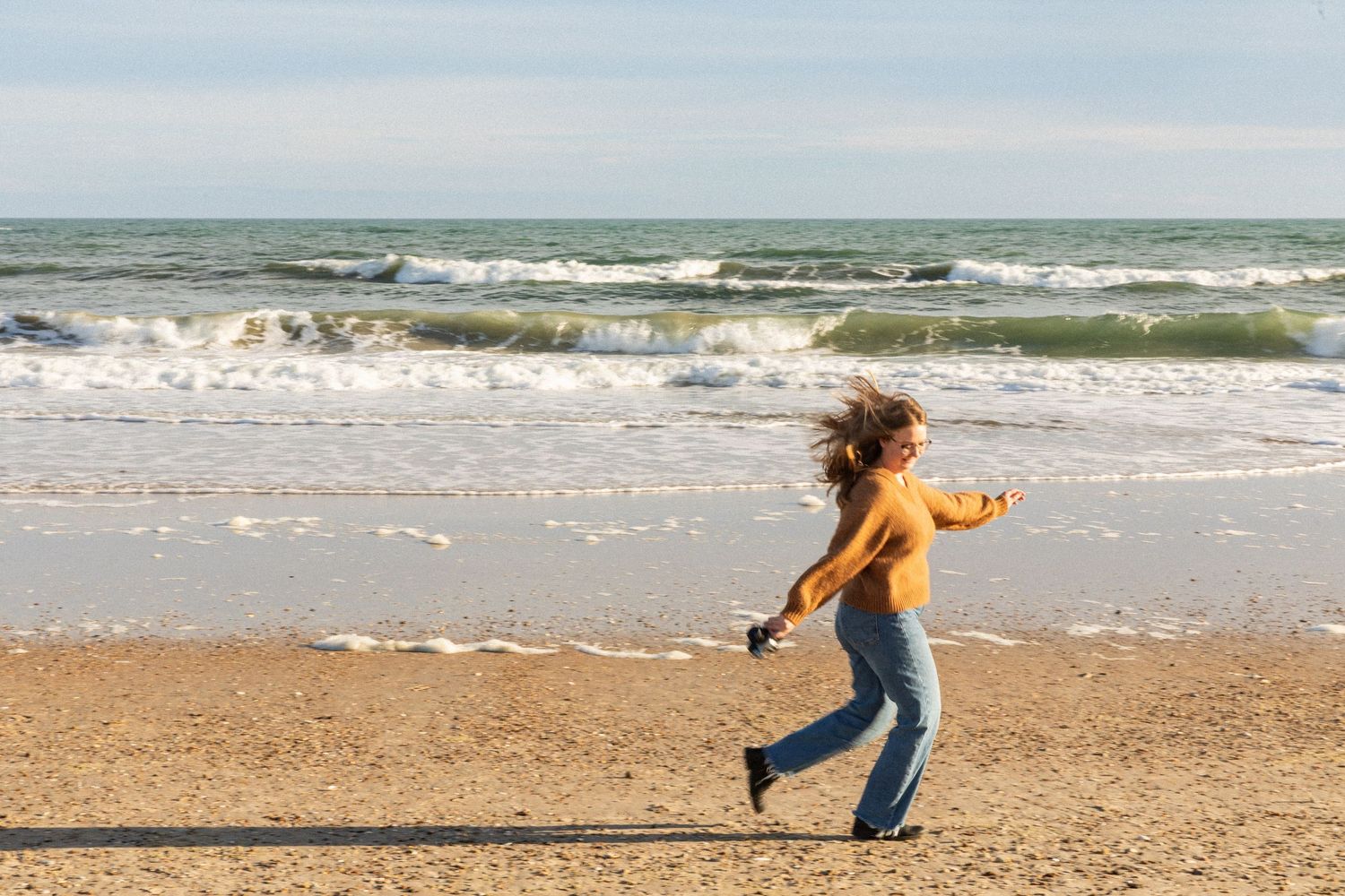 A joyful woman in a yellow sweater and jeans dancing on a sandy beach with ocean waves in the background in Jacksonville North Carolina