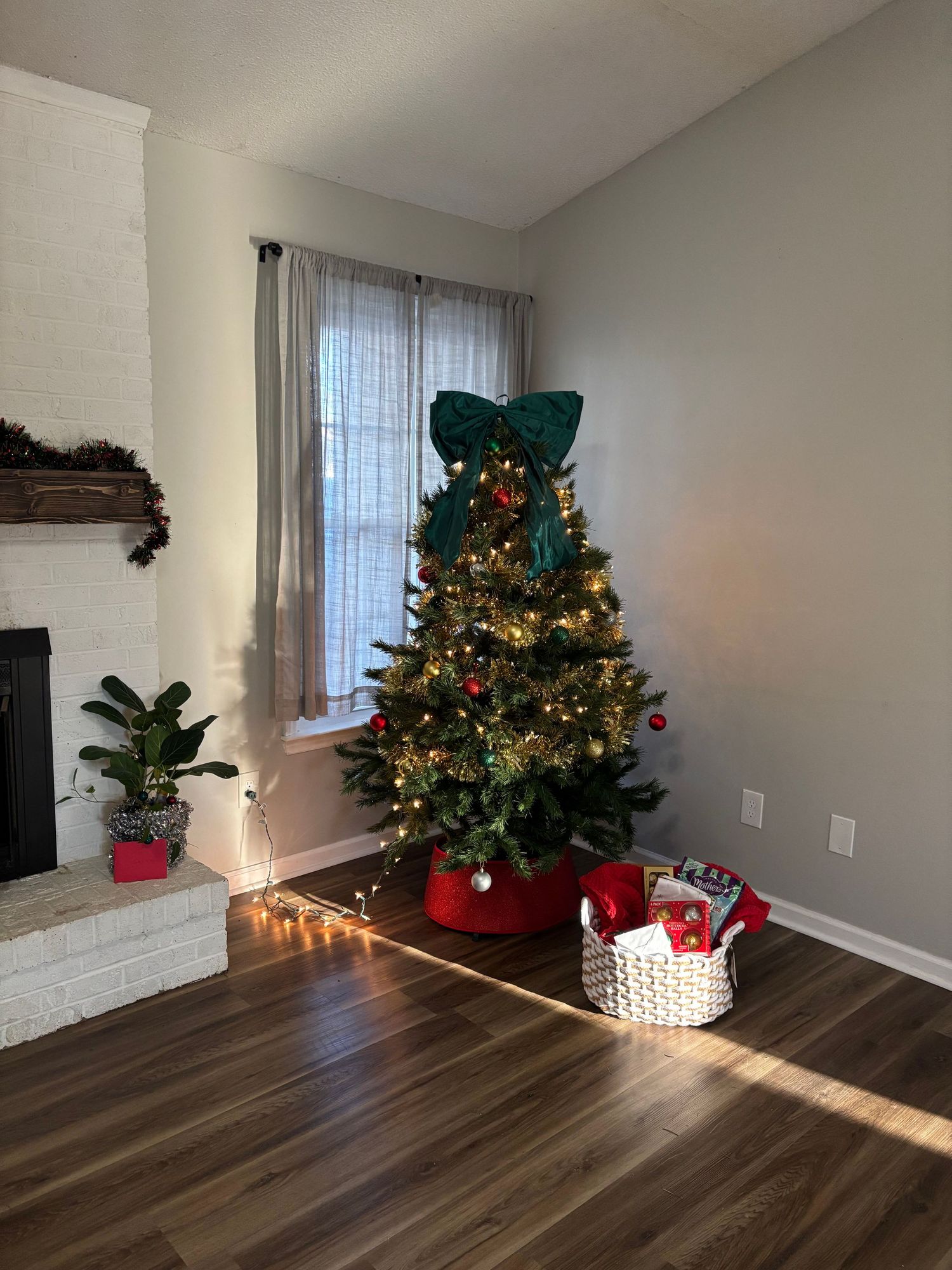 A decorated Christmas tree with warm lights and a green bow topper stands in a cozy living room corner with gifts nearby.