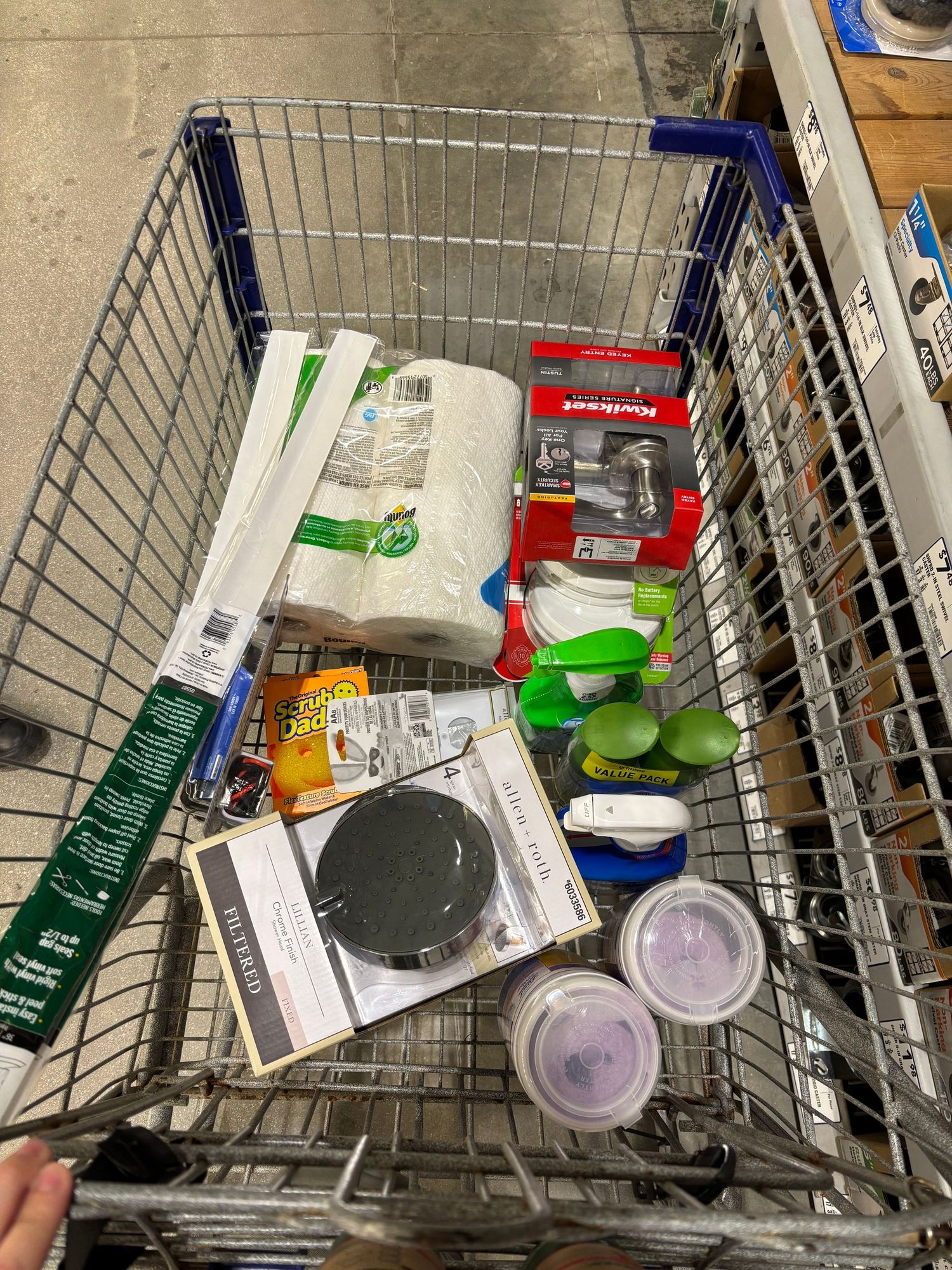 Shopping cart filled with household items including paper towels, storage containers, and kitchen supplies at a retail store.
