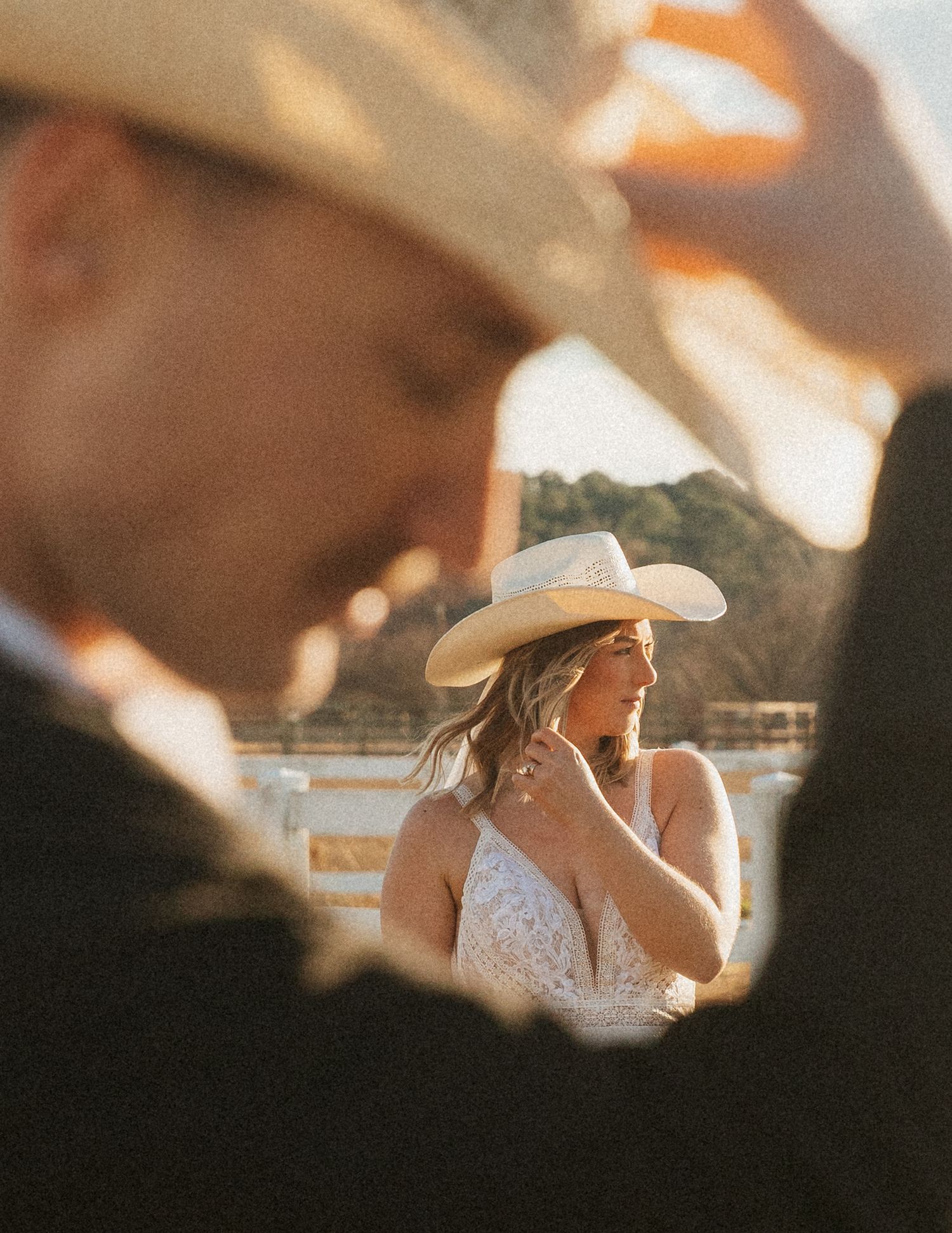 NC elopement. Bride in white lace dress and cowboy hat smiles softly at golden hour during rustic outdoor wedding ceremony.