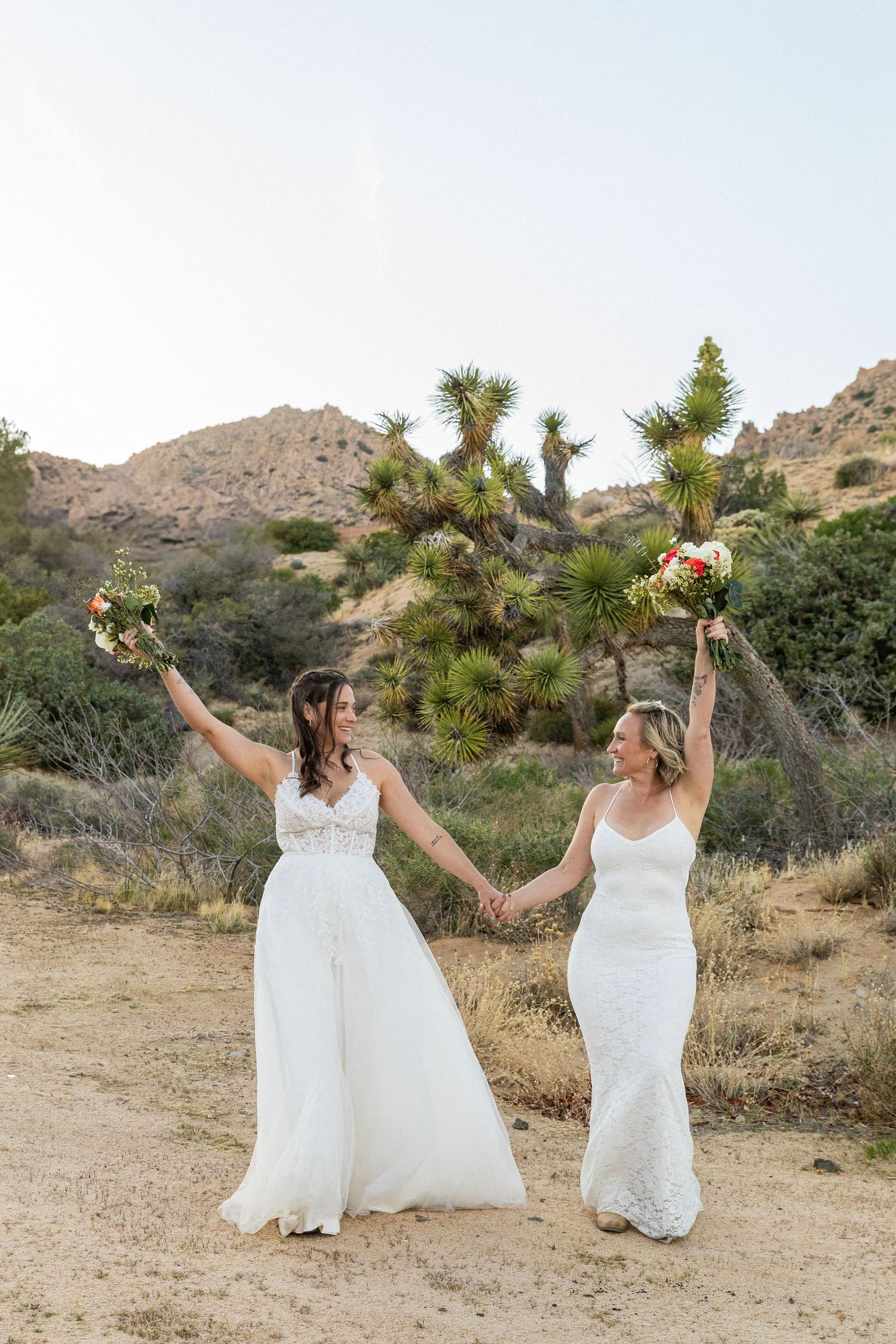 Two brides in white wedding dresses hold hands and raise bouquets while celebrating in a desert landscape with Joshua trees.