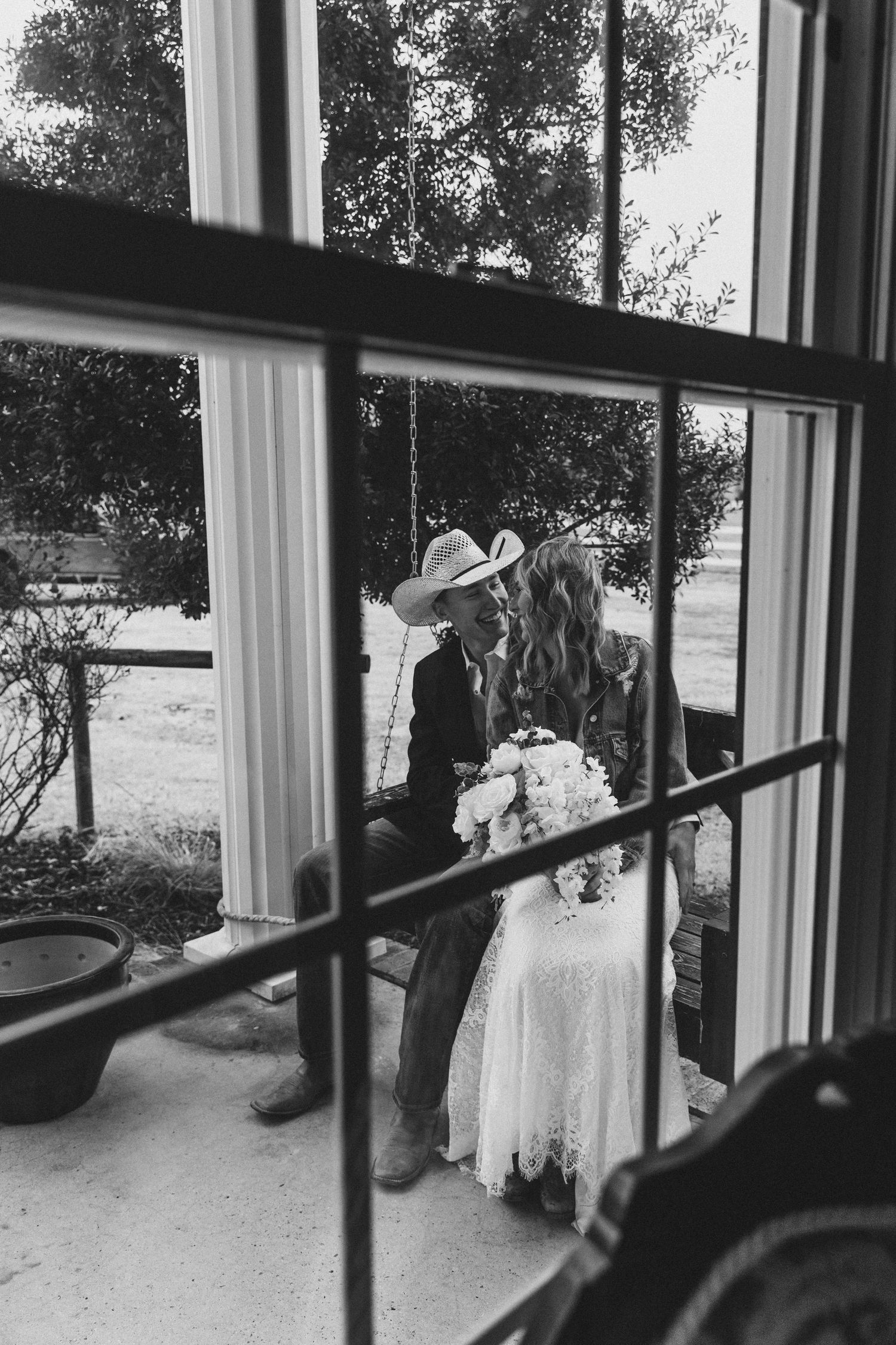 Black and white photo of a bride and groom sharing a kiss, viewed through a window, groom wearing a cowboy hat.