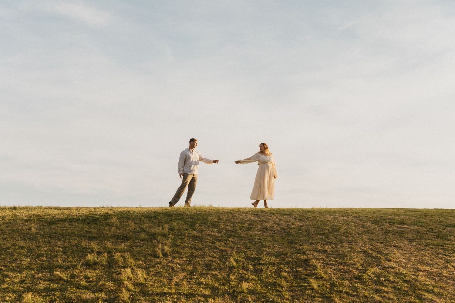Couple reaching toward each other on a grassy hill under a blue sky in a romantic outdoor setting.