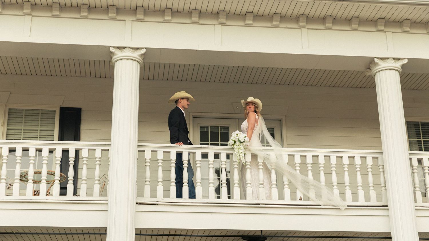Bride and groom in western attire standing on a white colonial-style venue balcony during an outdoor wedding ceremony.