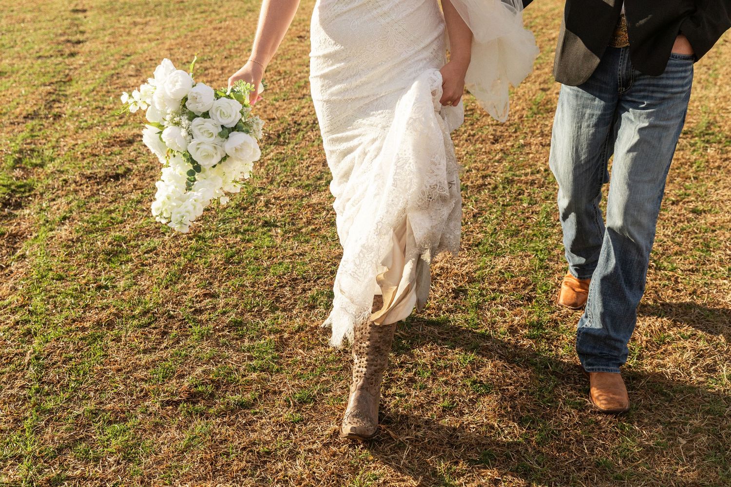 Bride in white wedding dress and cowboy boots holding bouquet walks with groom in jeans on green grass.