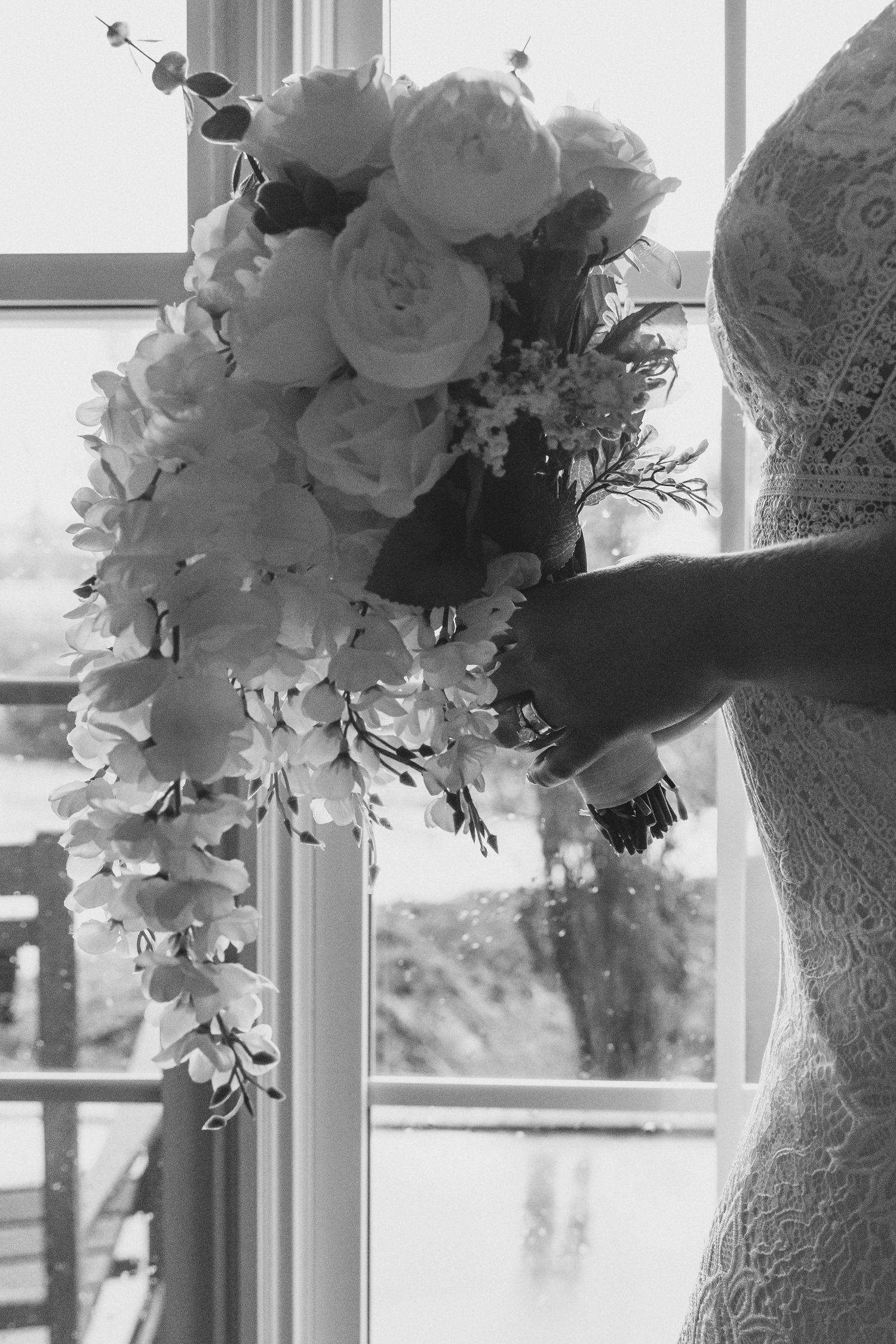 Black and white photo of a bride holding a cascading wedding bouquet of roses and orchids near a window.