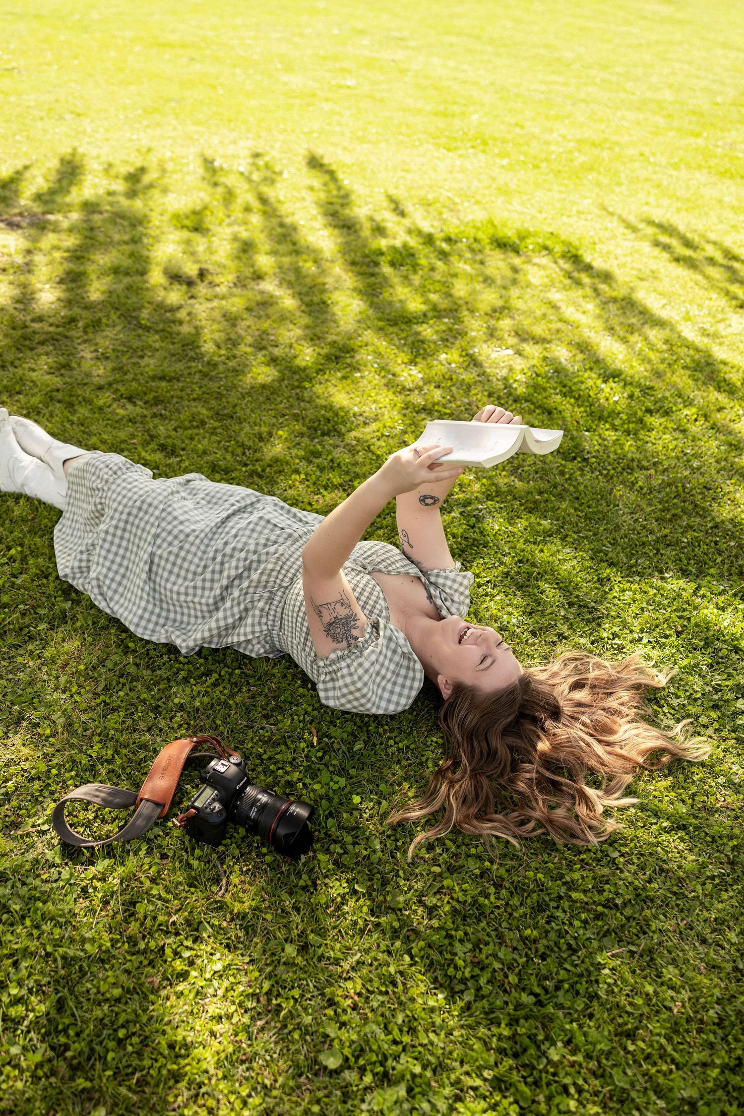 Woman in plaid dress lying on green grass, reading a book beside a camera on a sunny day in a park in Jacksonville North Carolina