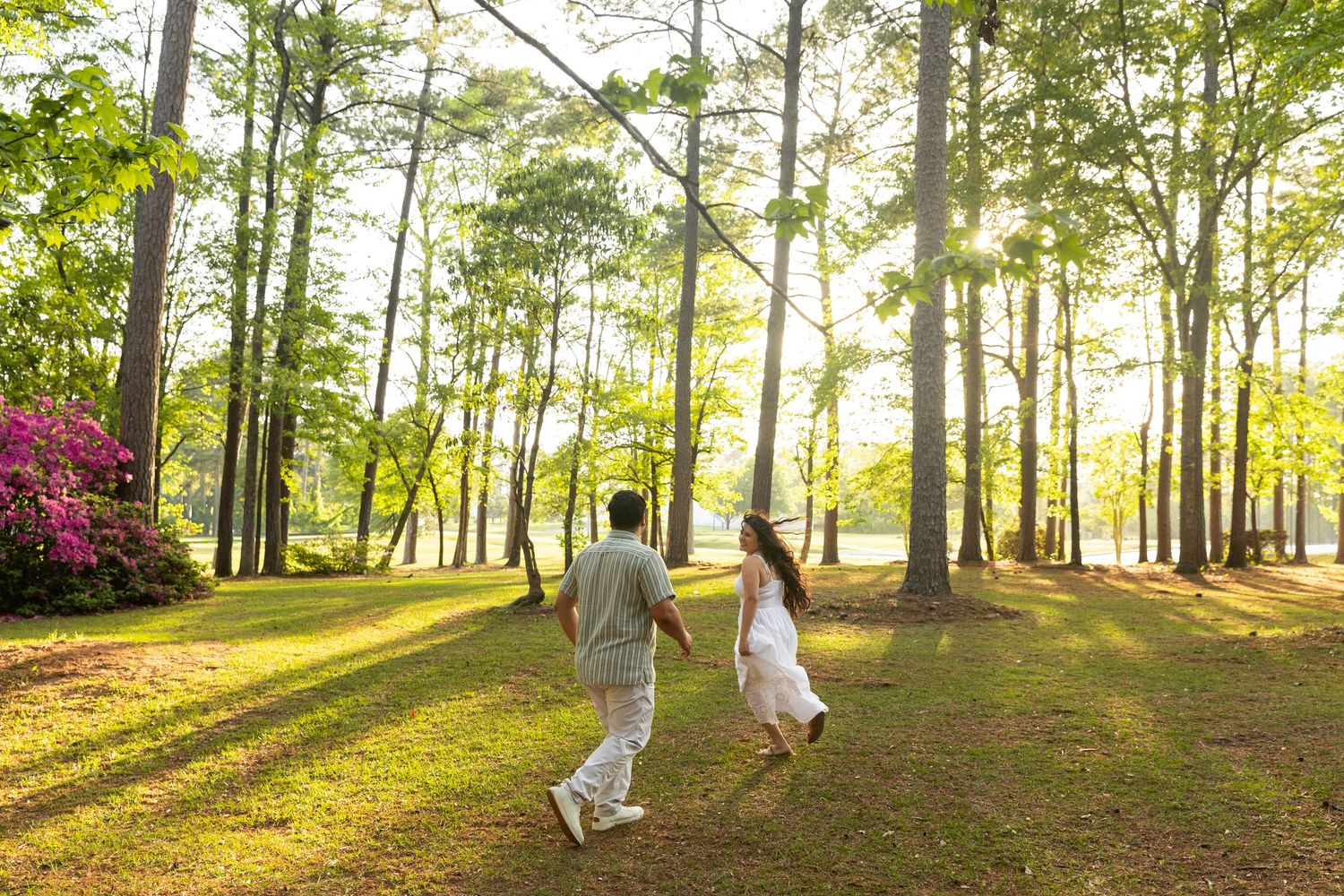 Couple holding hands running through sunlit forest with tall trees and green grass during golden hour in NC park