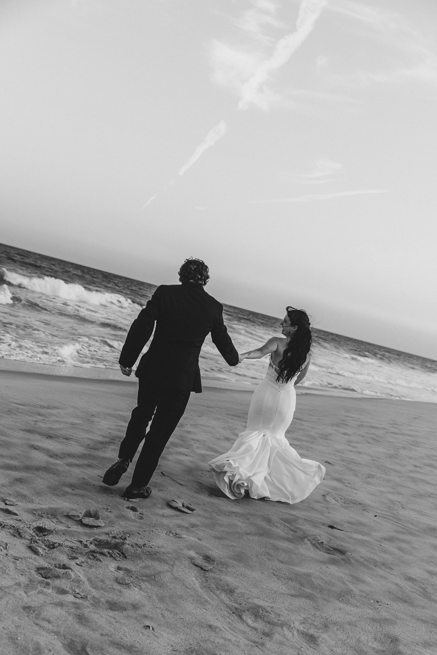 Black and white photo of a bride and groom holding hands, running along a sandy beach with ocean waves behind them.