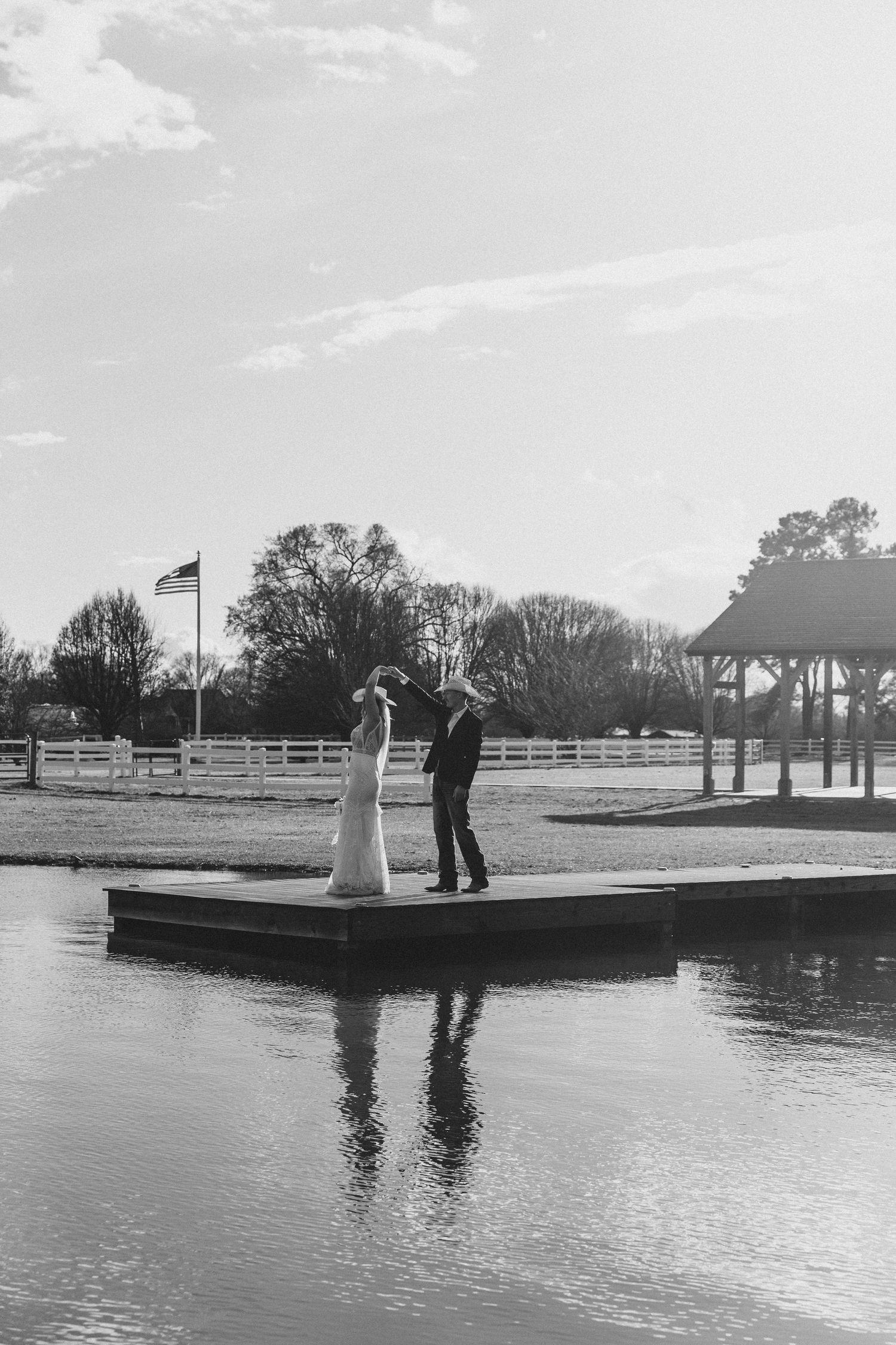 Black and white photo of a bride and groom standing on a dock over water at an outdoor wedding venue.