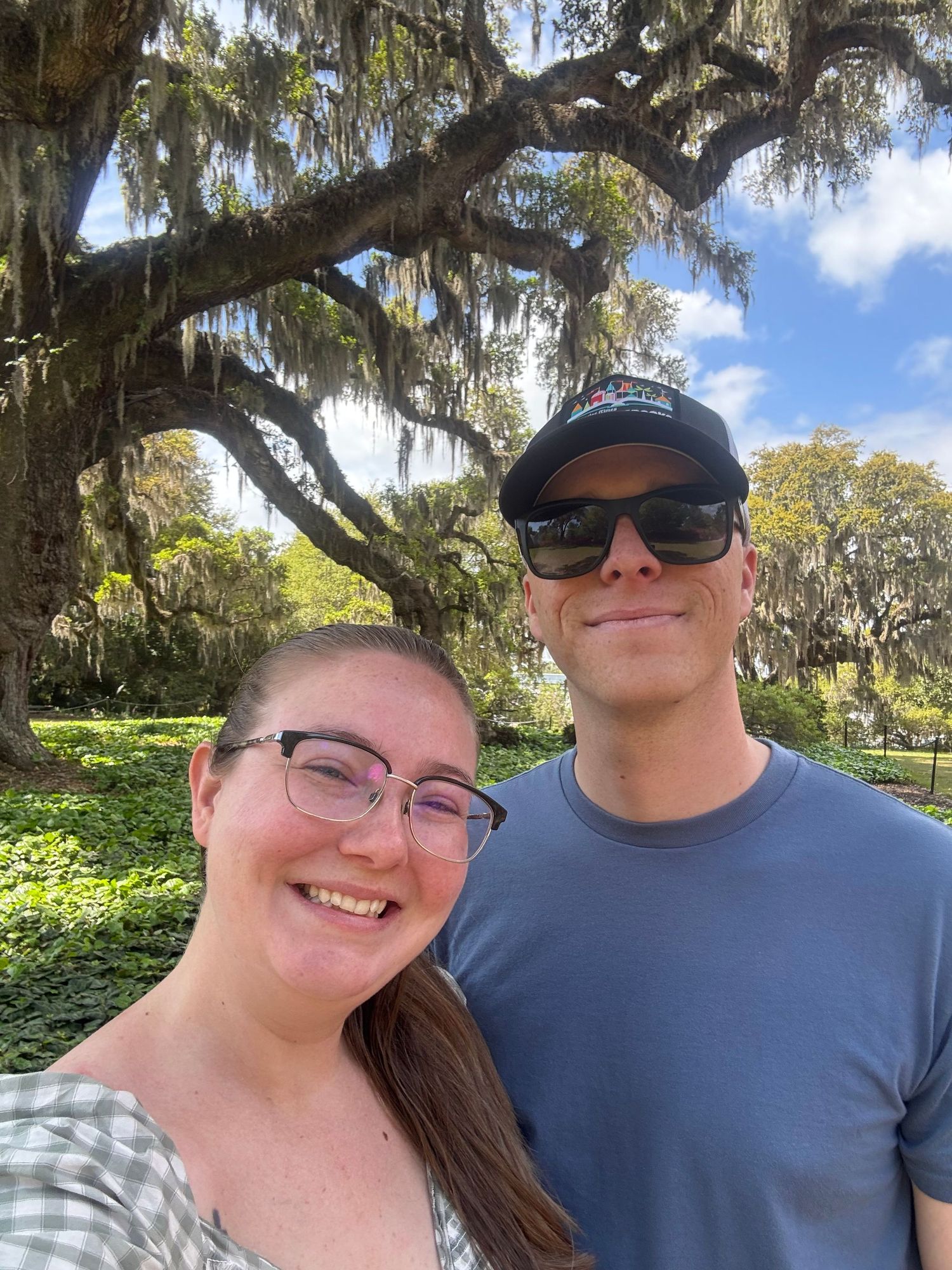 A smiling couple takes a selfie outdoors beneath sprawling moss-draped oak trees in a lush green park on a sunny day.