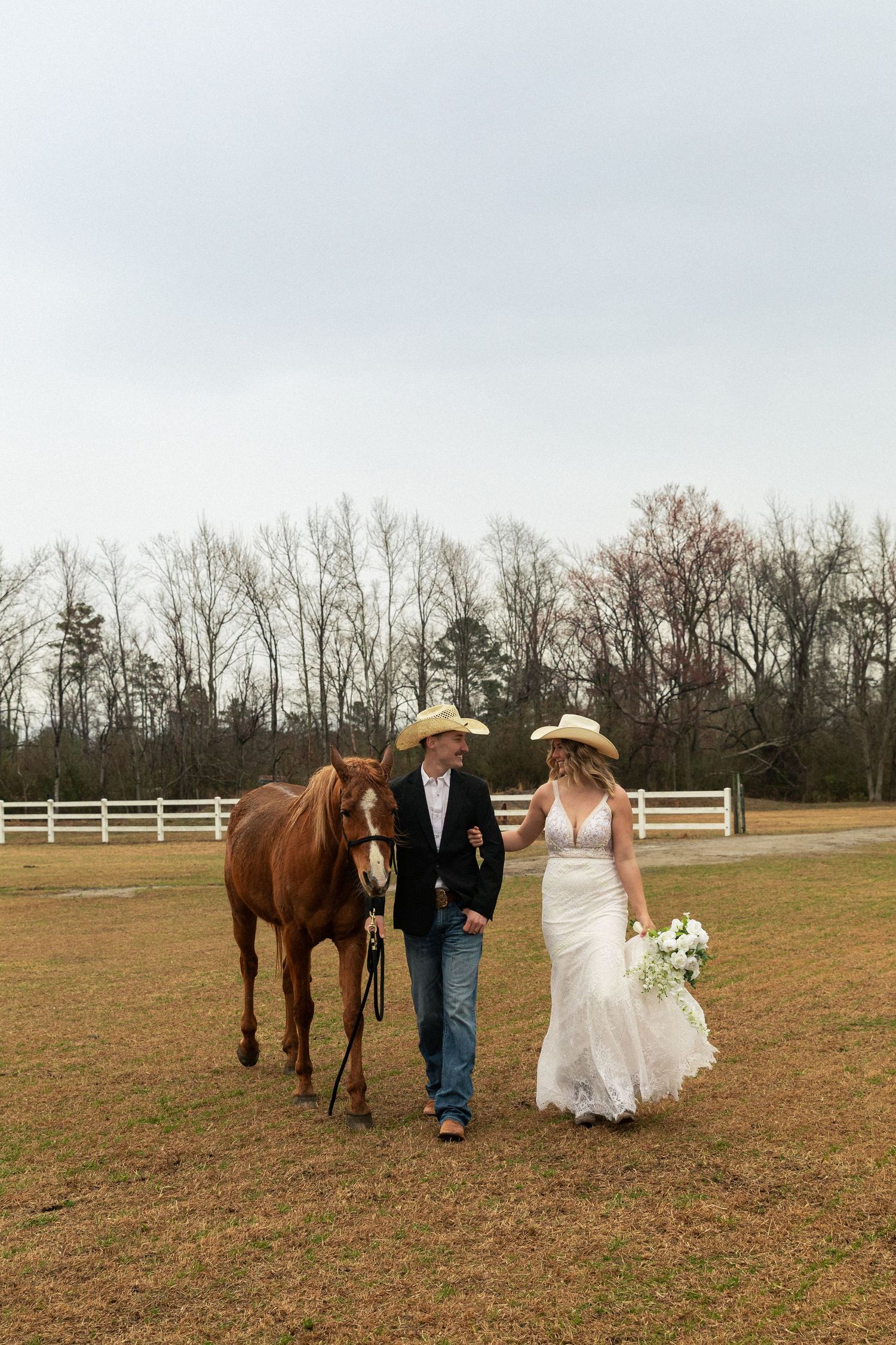 Bride and groom in cowboy hats walk with a chestnut horse on a farm, bride holding white bouquet in lace gown.