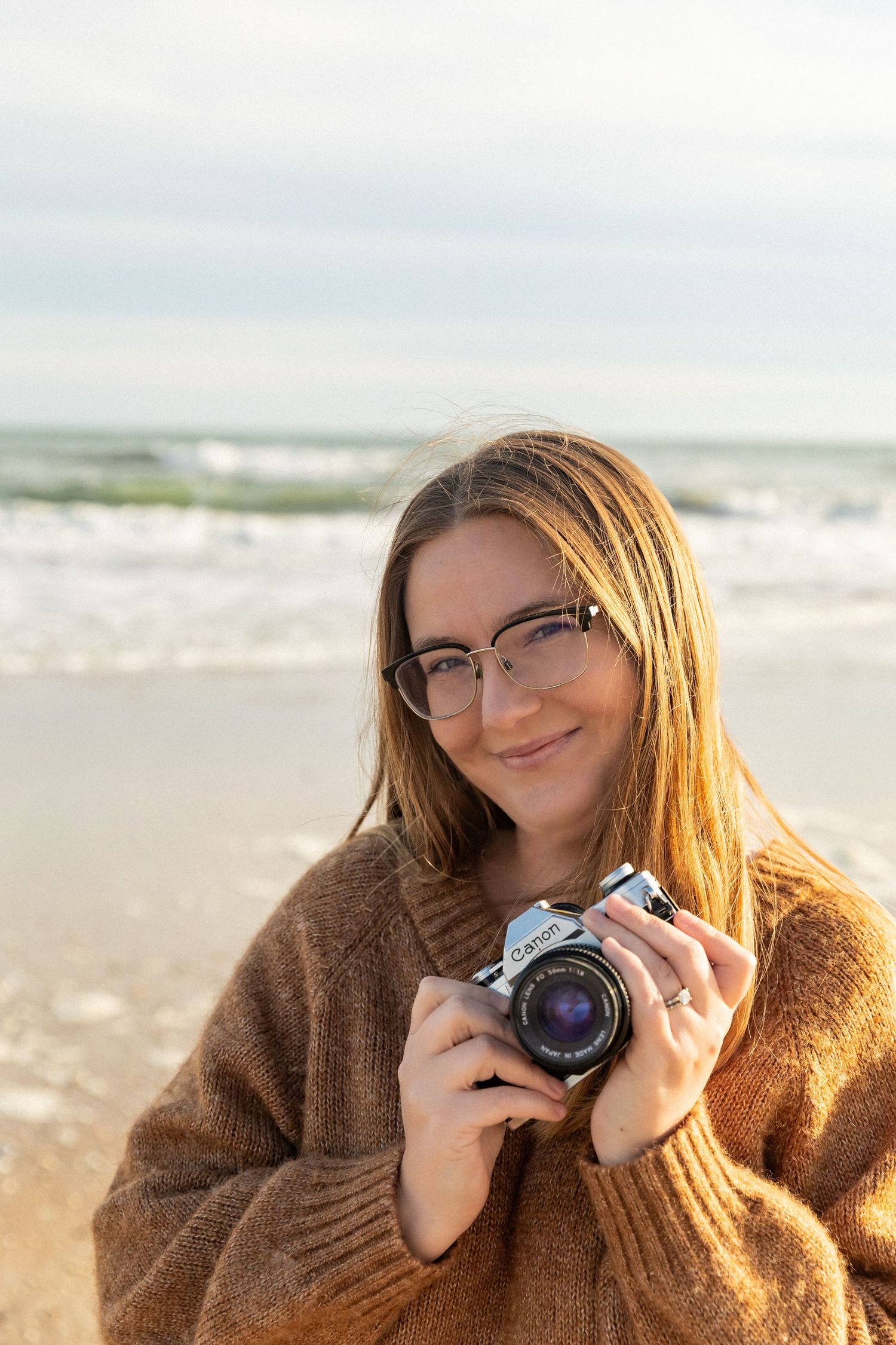 A photographer in a cozy brown sweater holds a camera while enjoying a sunny day at a beach in North Carolina.