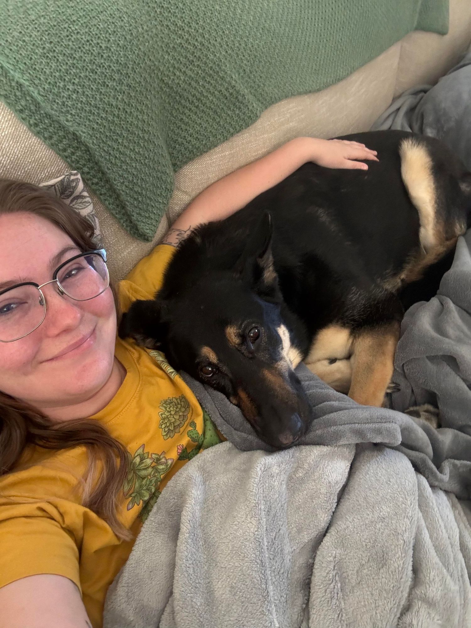 A young woman with glasses in a yellow shirt smiles while cuddling with a black and tan dog on a gray couch.