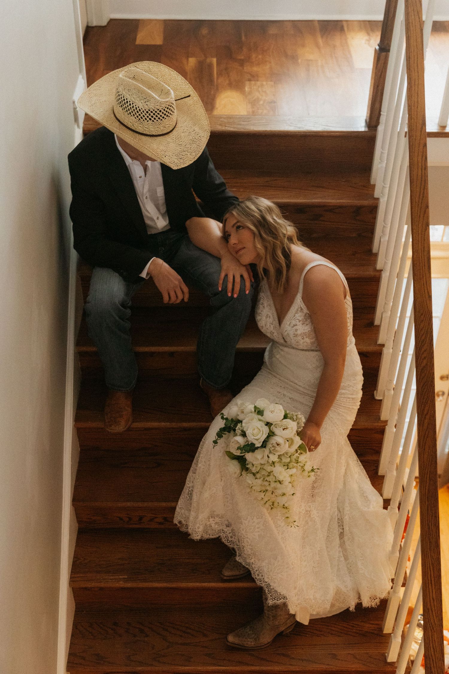 North Carolina Wedding photography. Bride in lace dress holding white bouquet sits on wooden stairs beside groom in cowboy hat and blazer.