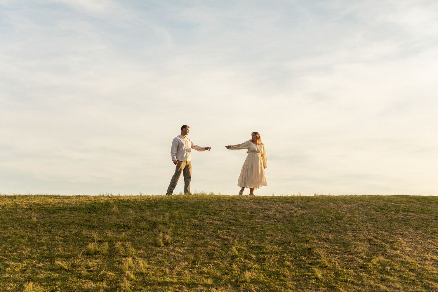 Couple reaching toward each other on a grassy hill at golden hour in an open field in Jacksonville North Carolina Park