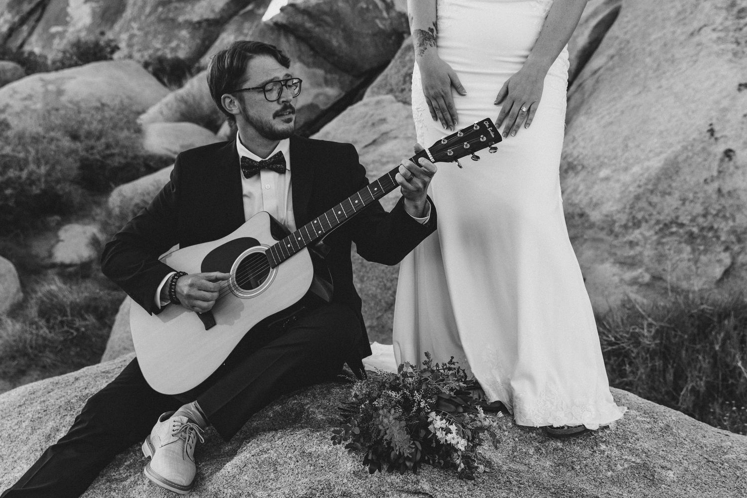 Groom in tuxedo playing acoustic guitar while bride in white dress stands nearby on rocks in black and white wedding photo.