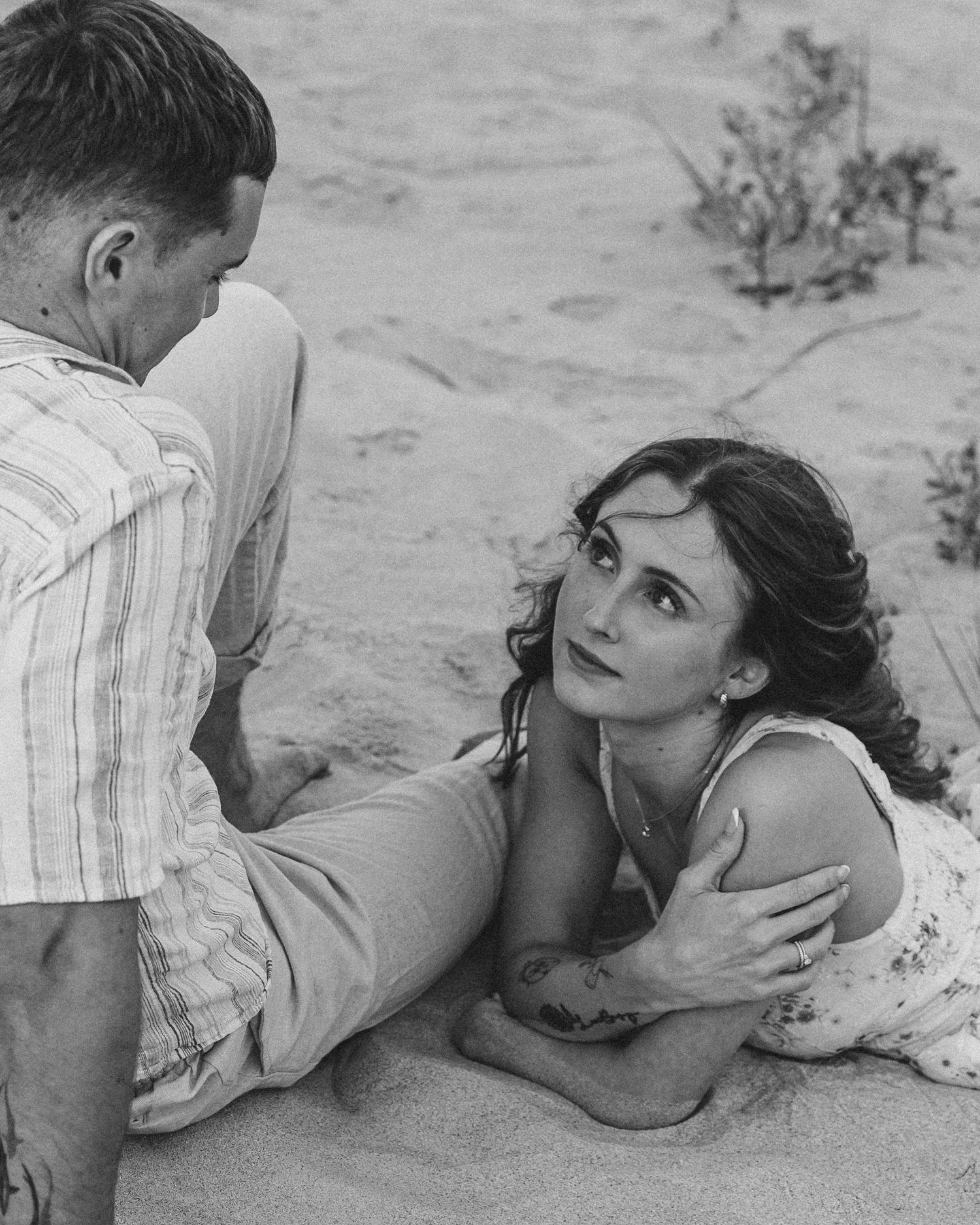 Black and white photo of a couple sitting on sandy beach, woman looking up at man romantically.