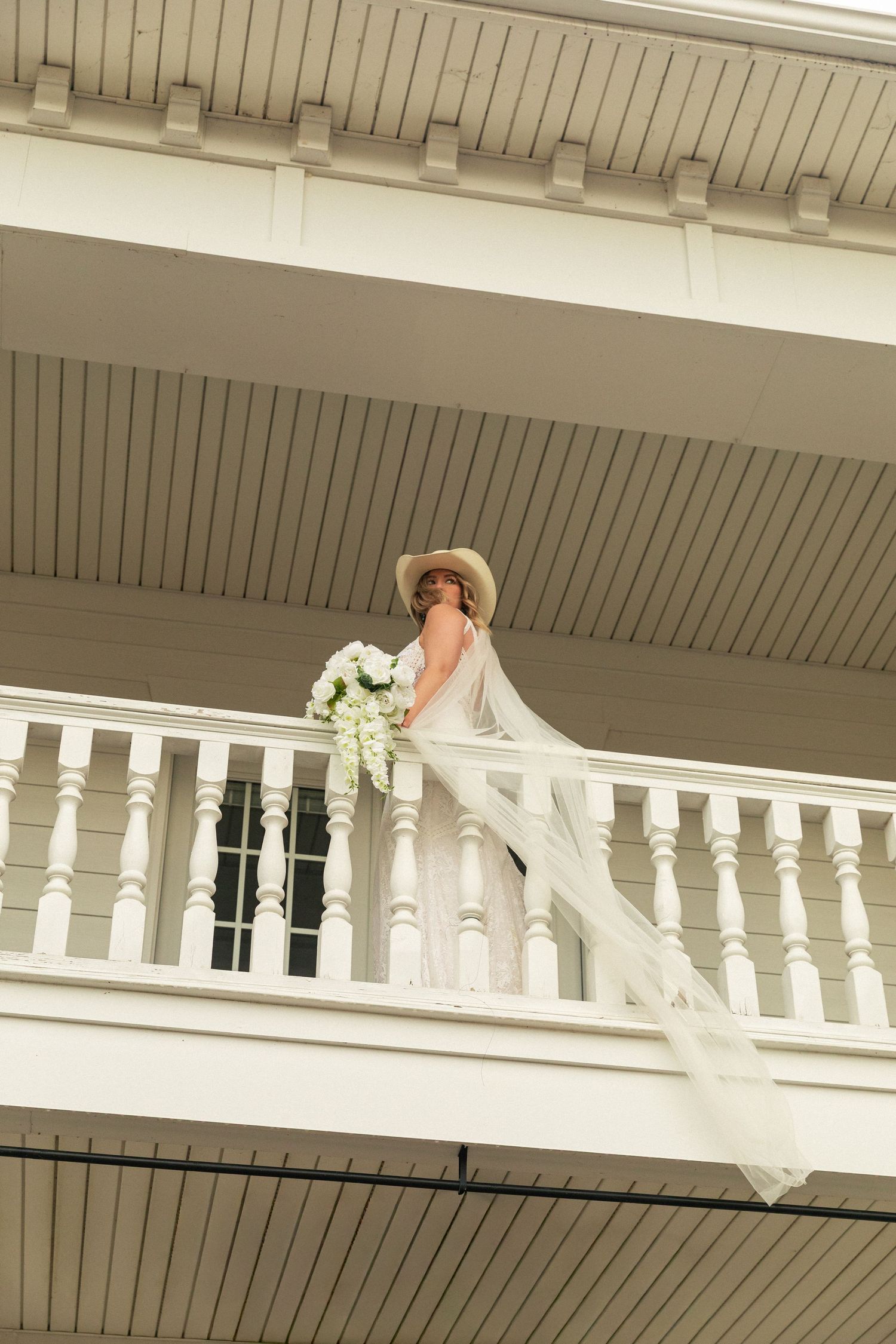 NC elopement photographer, Bride in white dress and cowgirl hat holds floral bouquet on white balcony with veil flowing in the wind