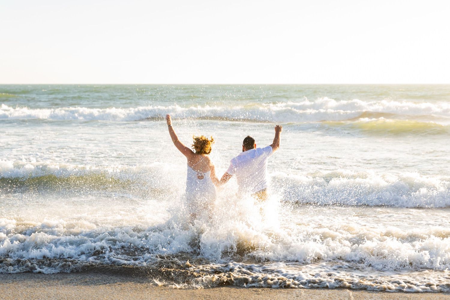 Coastal North Carolina Wedding. Couple celebrating in ocean waves on a sunny beach, splashing water with arms raised in joy