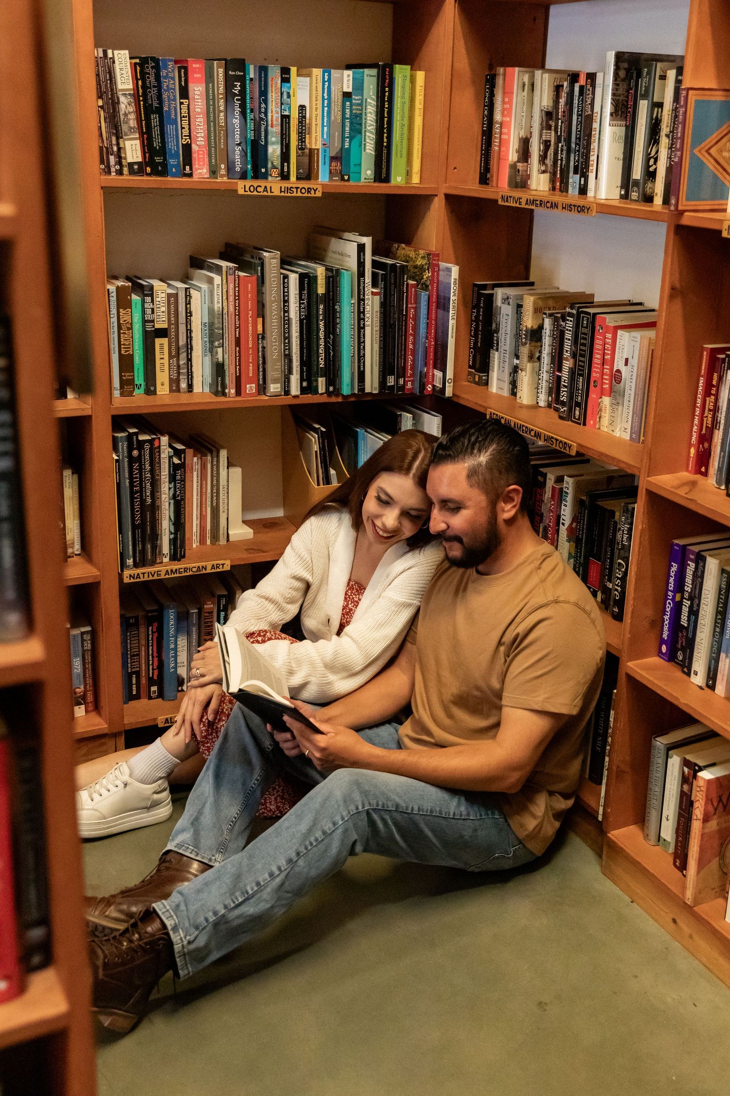Couple sitting on floor of cozy bookstore, sharing a book together among wooden shelves filled with colorful books.