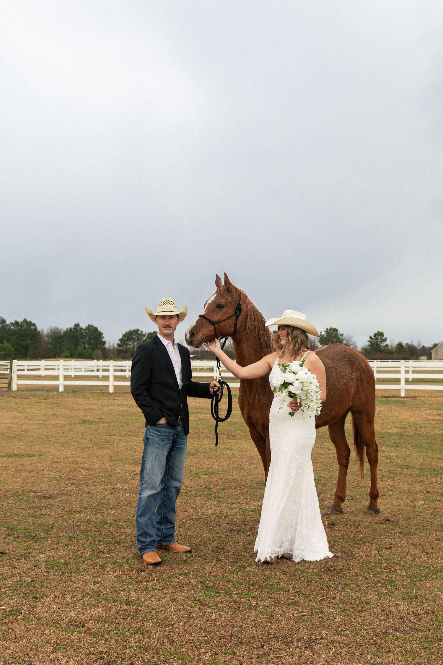 Bride and groom in cowboy hats pose with a chestnut horse at a rustic ranch wedding venue in Fayetteville North Carolina.