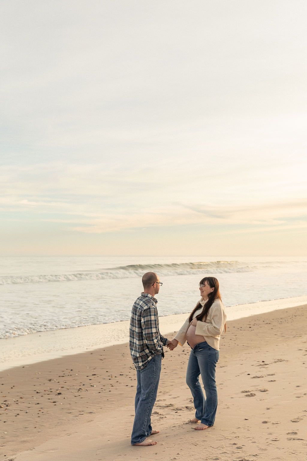 Pregnant couple holding hands on a sandy beach at sunset, waves gently rolling in behind them.