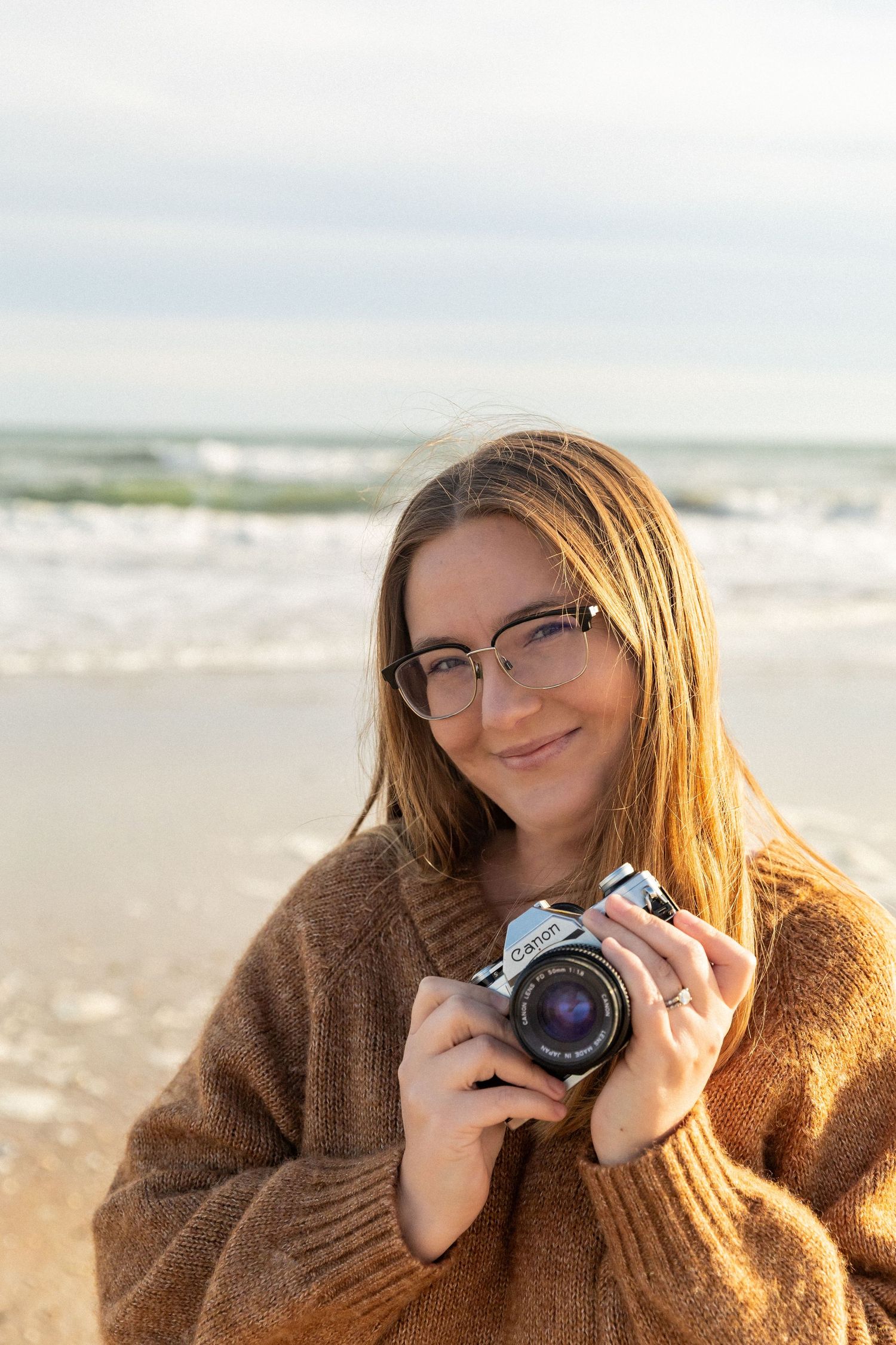 North Carolina Photographer with glasses holding a vintage Canon camera smiling at the beach in a cozy brown sweater.