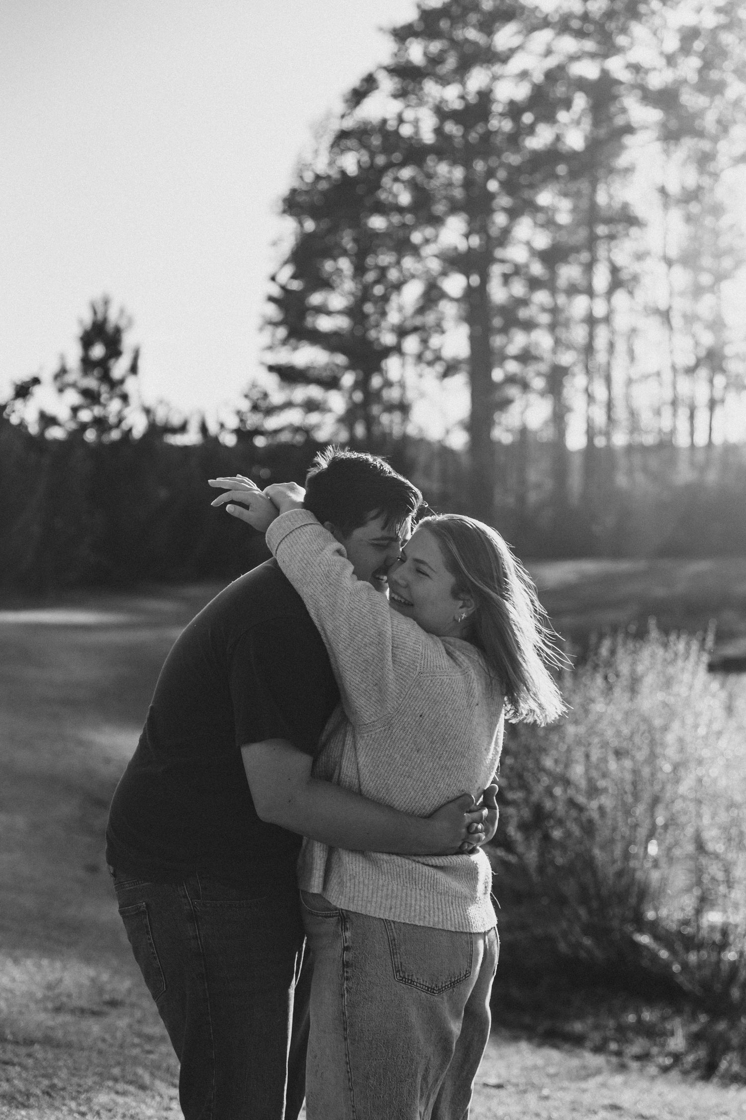 Black and white photo of a couple embracing and kissing outdoors on a path surrounded by trees.