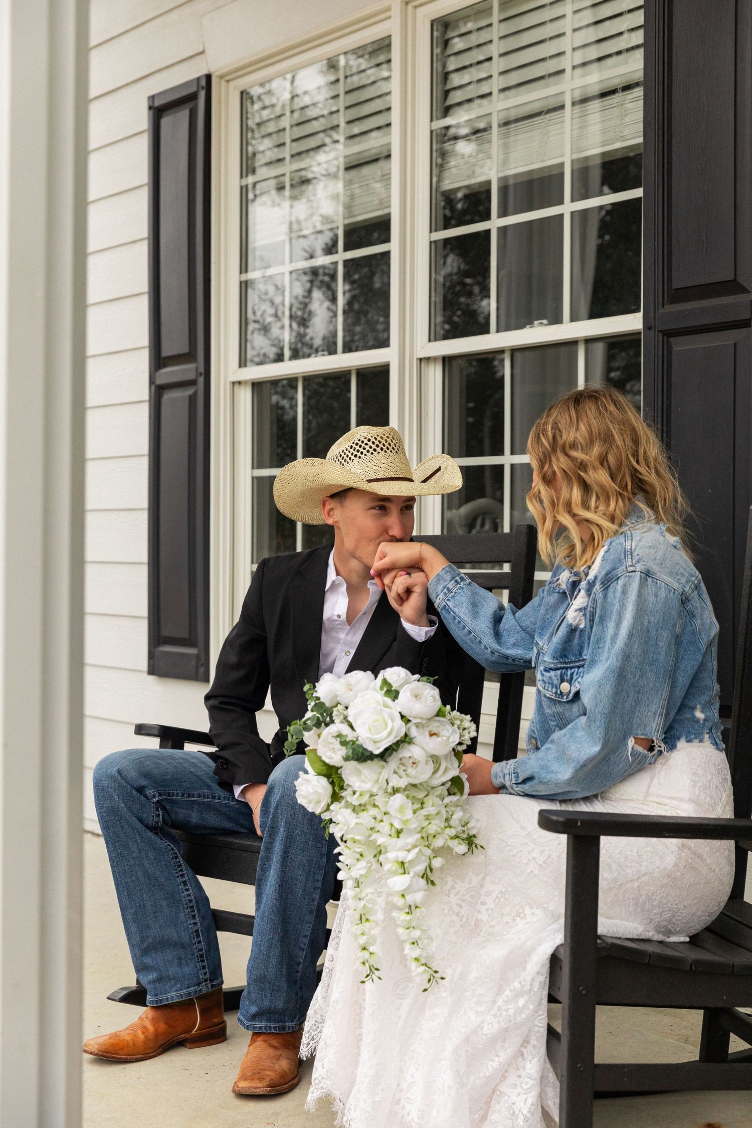 North Carolina wedding photo. Groom in cowboy hat kissing bride's hand on porch, bride wearing denim jacket holding white floral bouquet.