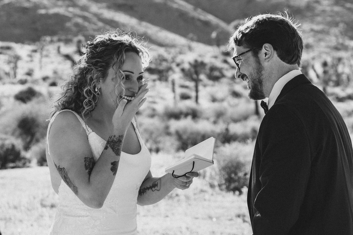 Black and white photo of a bride with tattoos reading wedding vows to her groom outdoors in a desert landscape.