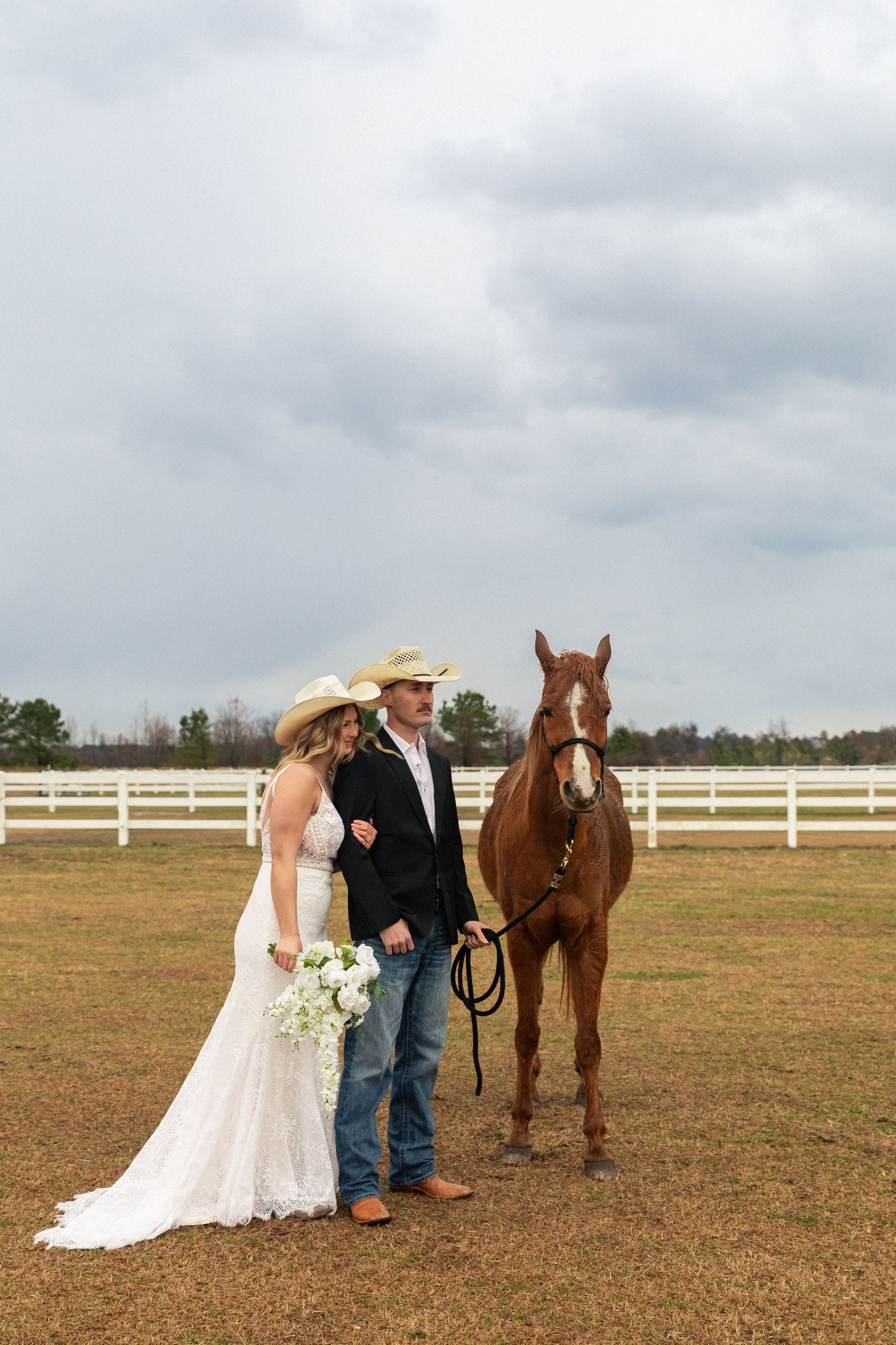 Bride and groom in cowboy hats pose with a chestnut horse by a white fence at their country wedding.