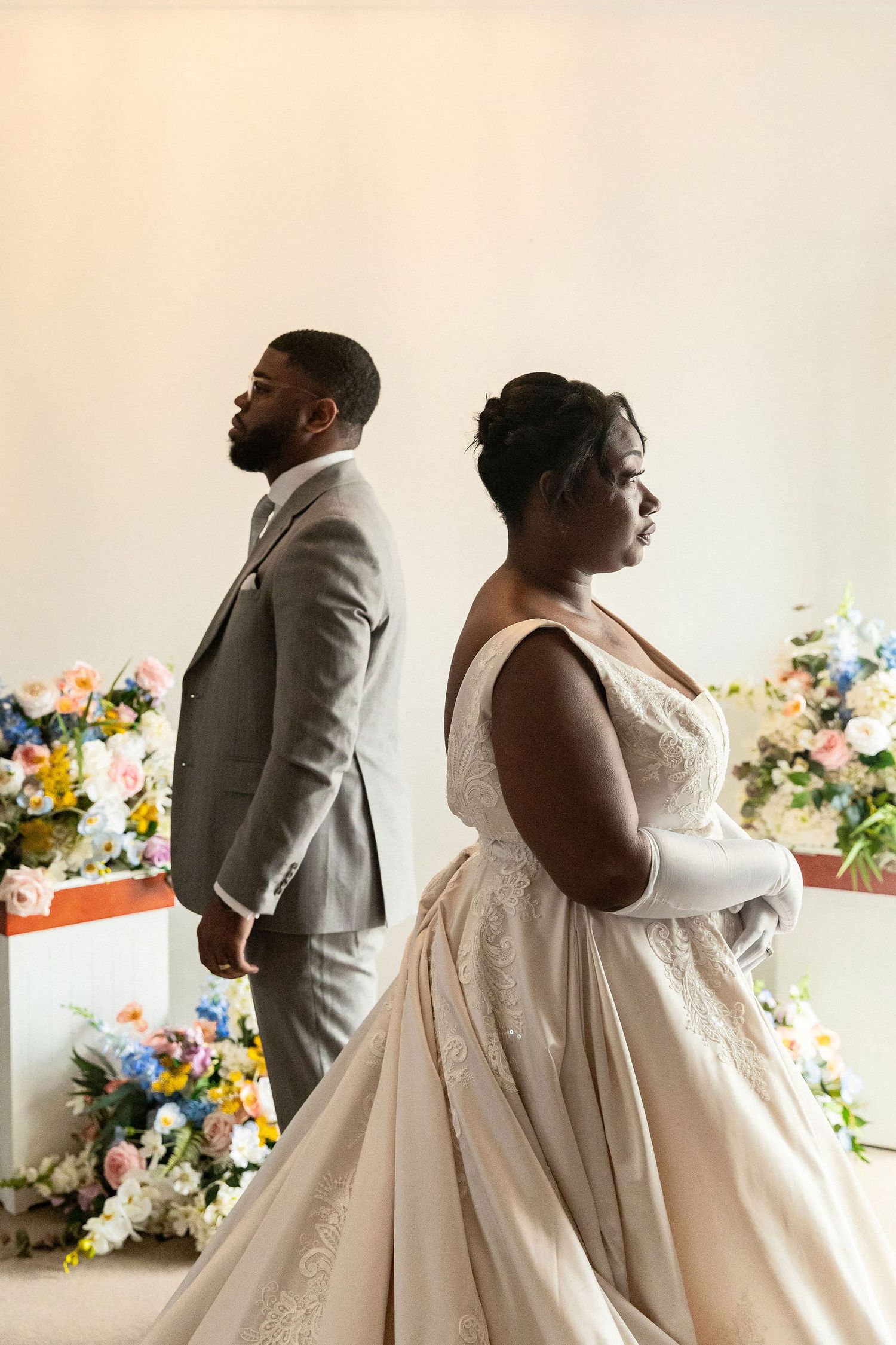 A bride in a lace ballgown and groom in a gray suit stand back-to-back at a floral-decorated wedding altar.