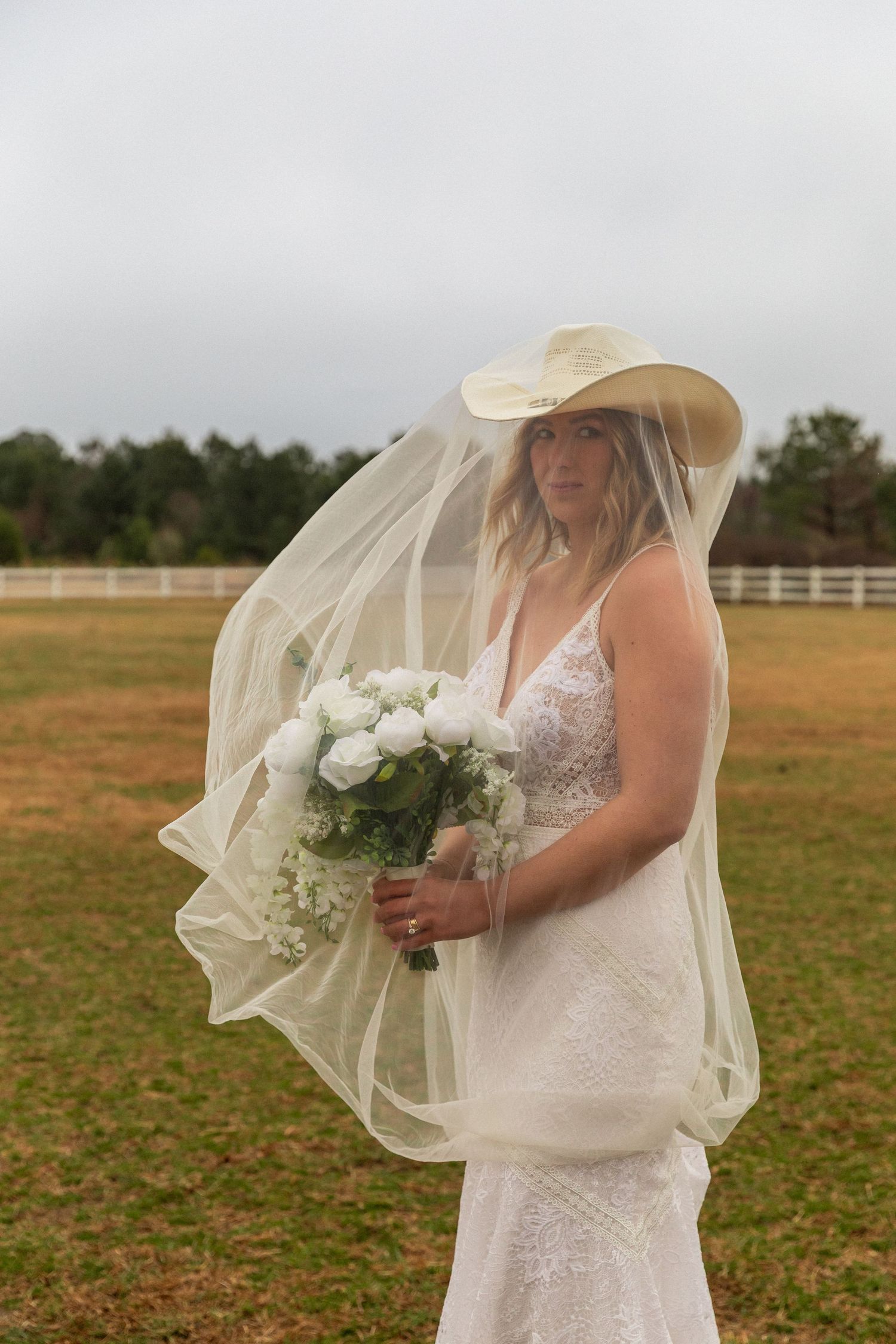 Bride in white lace gown and cowboy hat with veil holds white bouquet outdoors on green field in Eastern North Carolina